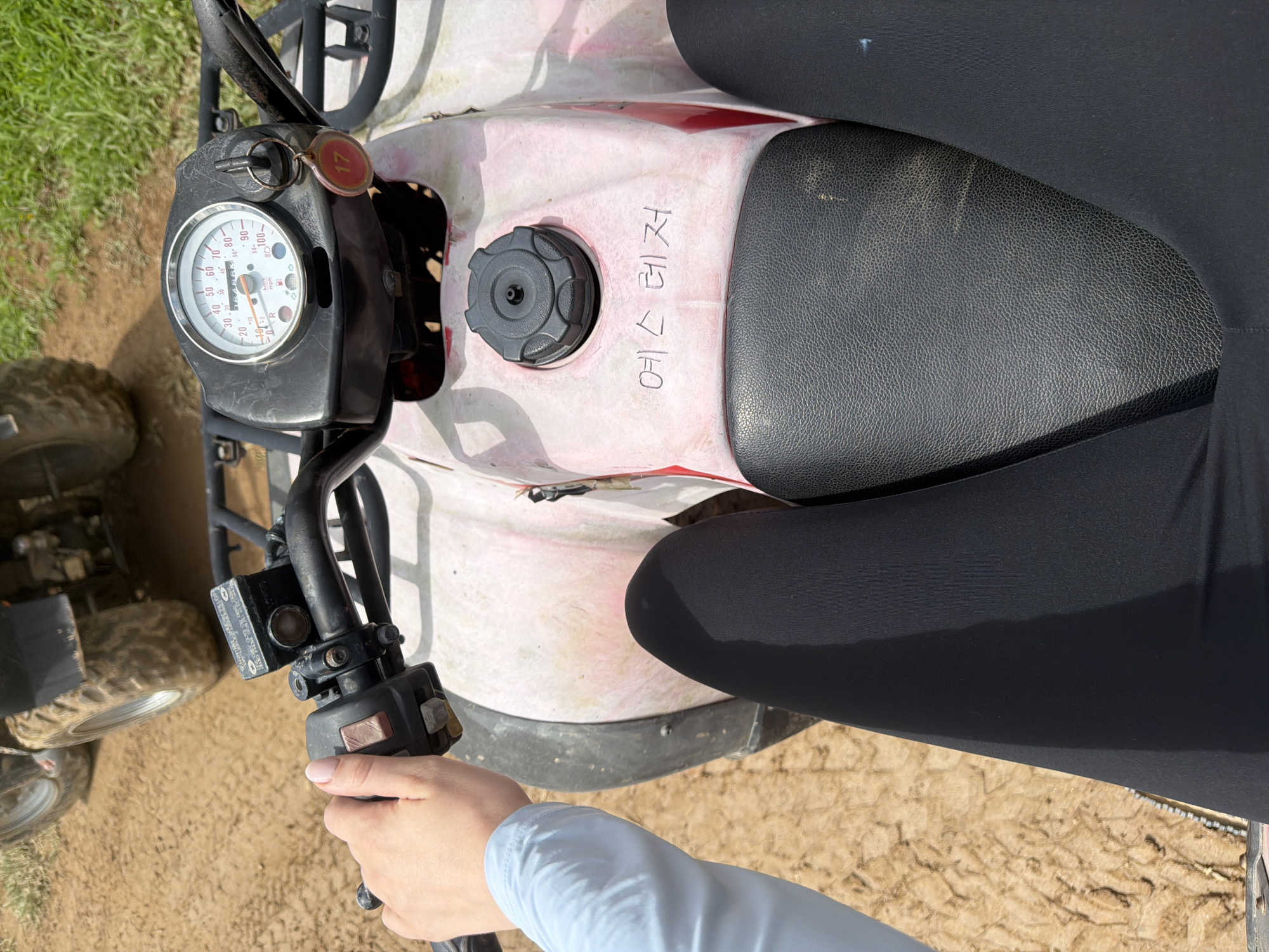A first-person perspective shows a person sitting on a pink and white ATV, holding the handlebars. The ATV has a speedometer displaying approximately 40 mph, a key in the ignition with a red tag labeled "17," and a fuel cap. Korean text, "예스 레저," is written on the ATV's body. The person is wearing a light blue long-sleeved shirt and black leggings. The background consists of a dirt path and green grass, suggesting an outdoor recreational area, possibly a leisure park in Seo-myeon, South Korea, given the presence of Korean writing. Another ATV is partially visible in the upper left corner of the image.