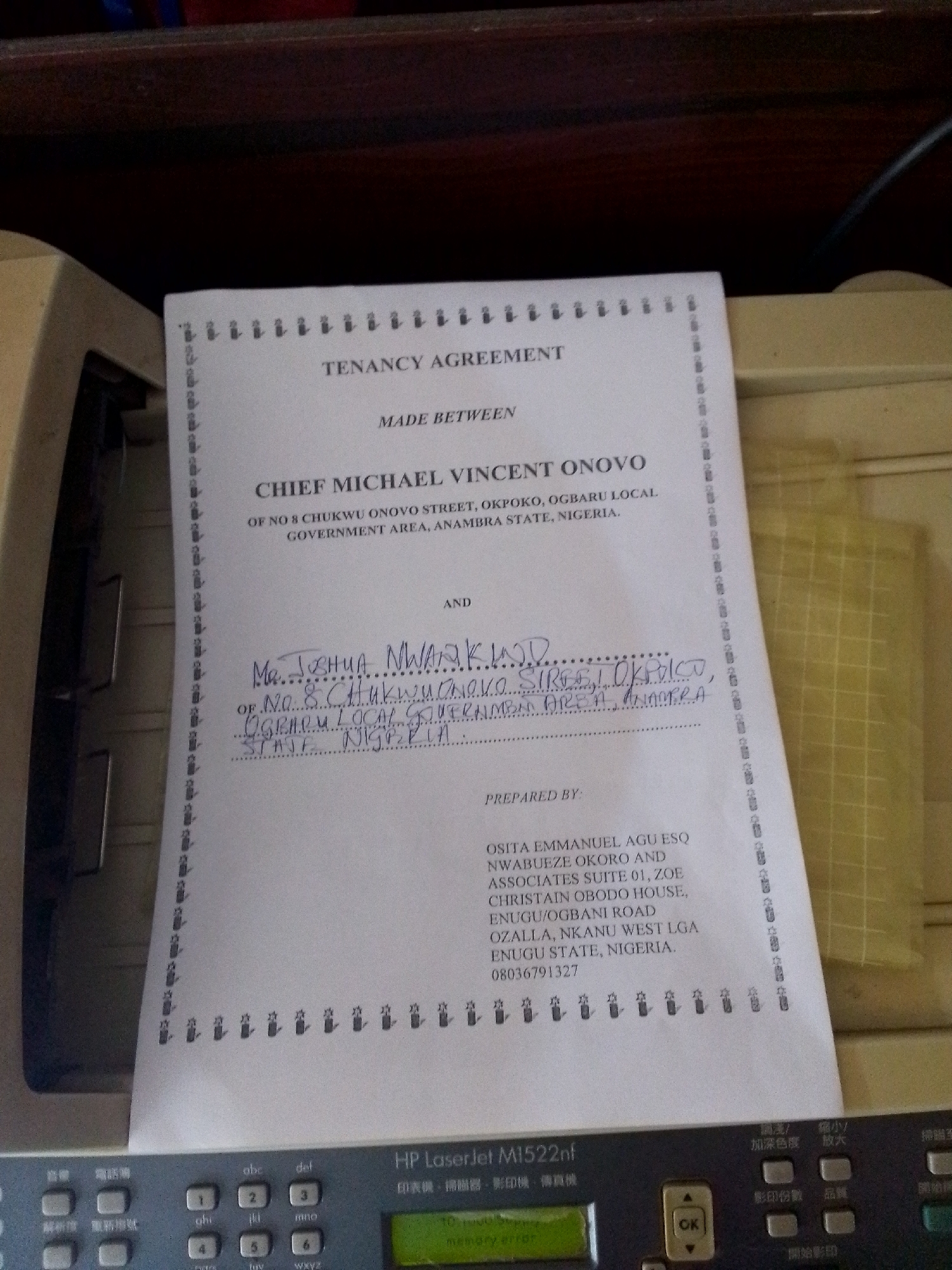 The image shows a close-up view of a document titled "TENANCY AGREEMENT". The document is printed on white paper with decorative borders and appears to be filled out by hand in places. The printed text identifies one party as "CHIEF MICHAEL VINCENT ONOVO" of "NO 8 CHUKWU ONOVO STREET, OKPOKO, OGBARU LOCAL GOVERNMENT AREA, ANAMBRA STATE, NIGERIA". The other party is identified by handwriting as "Mr JOSHUA MWAKUM D" of "NO. 8 CHUKWU ONOVO STREET, OKPOKO, OGBARU LOCAL GOVERNMENT AREA, ANAMBRA STATE, NIGERIA". The document also lists "OSITA EMMANUEL AGU ESQ NWABUEZE OKORO AND ASSOCIATES SUITE 01, ZOE CHRISTAIN OBODO HOUSE, EZUGU/OGBANI ROAD, OZALLA, NKANU WEST LGA ENUGU STATE, NIGERIA. 08036791327" as the preparers. The document is placed on top of a printer. The printer is a Hewlett-Packard Laserjet M1522nf. The environment appears to be indoors. There are no people clearly visible in the image.