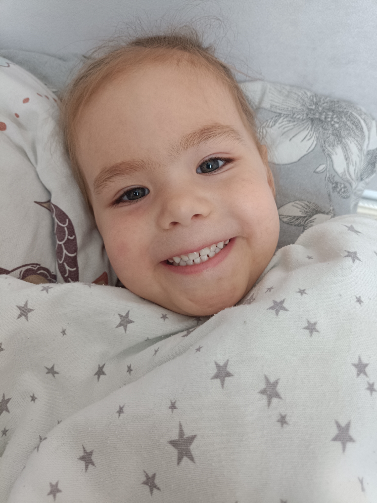 The image shows a close-up, overhead view of a young child lying in bed, wrapped in a white blanket with a pattern of grey stars. The child has fair hair, blue eyes, and is smiling broadly, revealing their teeth. The child is positioned on a pillow with a floral pattern and a blanket with a brown scale pattern on the left side. The background is out of focus. There is no indication of the location in Noviy, Russia.
