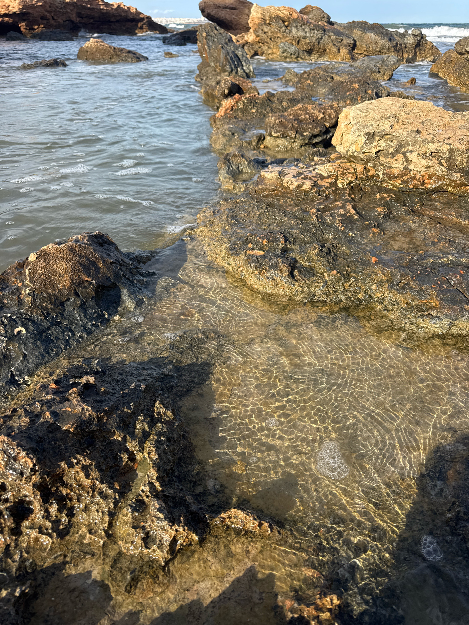 A low-angle, close-up shot captures a rocky coastline at the edge of the ocean. The foreground is dominated by dark, porous rocks with some patches of lighter brown and orange hues. The rocks are wet and glistening, with small pools of clear water collecting in crevices. The water's surface is textured with ripples and reflects the sunlight, creating a shimmering effect on the rocks below. Several small bubbles are visible in the shallow water. Further back, larger, more rugged rocks emerge from the sea. The ocean water appears murky grey-blue with some white foam near the shore. In the distance, the horizon is blurred, with a hint of land or a distant structure visible against a bright sky. The lighting suggests it is daytime, possibly late afternoon or early morning due to the warm tones on some of the rocks.