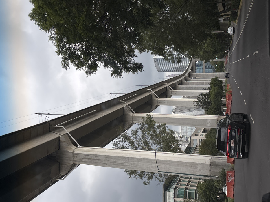 The image depicts an urban street scene in Mexico City, specifically along **Periférico Sur**, near the elevated viaduct of Metro Line 12 in the vicinity of Jardines del Pedregal or San Jerónimo.
The foreground shows a multi-lane asphalt roadway with dashed white lane markings. A dark-colored SUV, bearing license plate RUC-65-84, is observed from the rear, traveling away from the camera in the left lane. Orange plastic barriers are positioned along the left side of the road, appearing to delineate a median or construction area.
The dominant feature of the scene is a massive, elevated concrete viaduct that curves through the frame, supported by numerous tall, cylindrical concrete pillars. This structure carries an overhead metro line, evidenced by visible catenary wires. The underside of the viaduct deck features white, diagonal support braces connecting to the pillars.
On the far left, a modern multi-story building with green-tinted glass and light-colored panels is partially visible. Trees with green foliage are present along both sides of the road and between the elevated structure's pillars. In the background, to the right of the elevated structure, a tall, cylindrical building with a distinctive horizontally striped facade is prominently visible, bearing the name "POSADAS" near its summit. Other modern buildings are visible through the trees in the distance. The sky is overcast with gray clouds, indicating diffuse daylight. No pedestrians are visible.