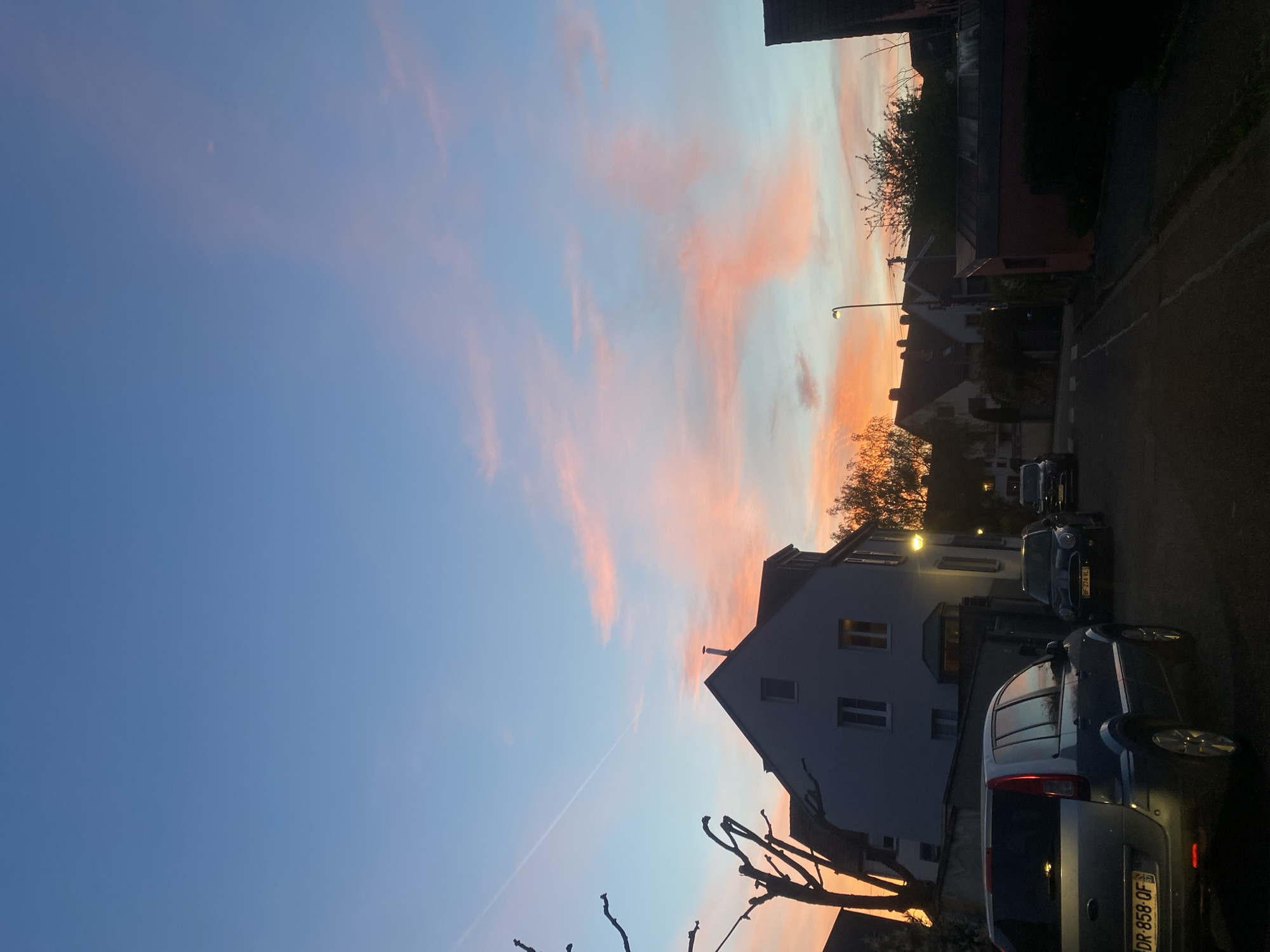 The image captures a street scene at dusk in Strasbourg, France. The sky is the dominant feature, transitioning from a deep blue to a soft orange-pink at the horizon. Wisps of clouds are illuminated with the setting sun's hues.
A two-story house is partially visible on the right side of the frame, its windows reflecting the diminishing light. Parked cars line the street, with a grey car in the foreground showing a license plate that reads "DR-858-QF". A dark asphalt road runs along the right edge of the frame. Silhouetted trees and building outlines are visible further down the street. A street lamp casts a faint glow.
There are no people visible in the image.