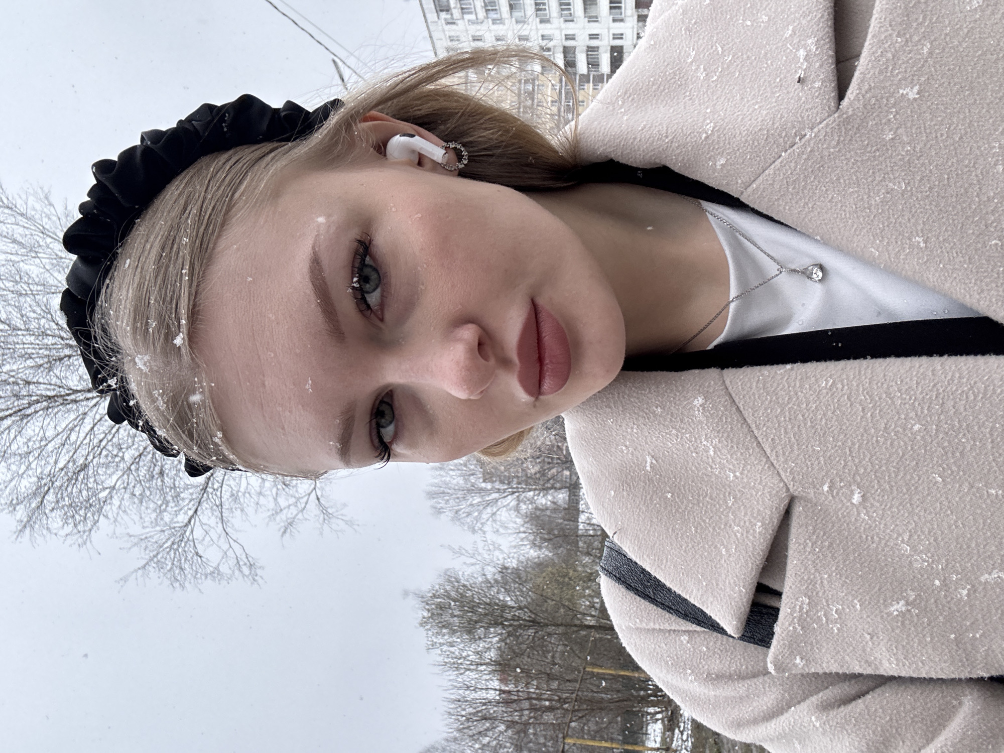 The image is a close-up, vertical outdoor portrait of a young woman, rotated 90 degrees counter-clockwise. She is looking directly at the camera with a neutral expression.
The subject is a young woman with light-colored hair (blonde/light brown) and light-colored eyes. Snowflakes are visible on her hair and face. She is wearing a black ruffled headband. In her right ear, a white Apple AirPods earbud is visible, along with a small, silver-toned circular earring. Her lips have a light pink tint.
She is dressed in a light beige or off-white textured coat, which is flecked with snowflakes. A black strap or trim is visible over her left shoulder. Beneath her coat, she wears a white top, showcasing a delicate silver-colored necklace with a single, clear teardrop-shaped gemstone pendant.
The background suggests an urban or suburban setting in Perm, Russia, during winter or early spring. Bare trees with fine branches are prominent in the middle ground against an overcast, light grey sky. A multi-story building, likely an apartment block, can be seen in the upper right background. Snowflakes are actively falling and have settled on the woman's clothing and hair, indicating cold, snowy weather. There is no visible text in the image.