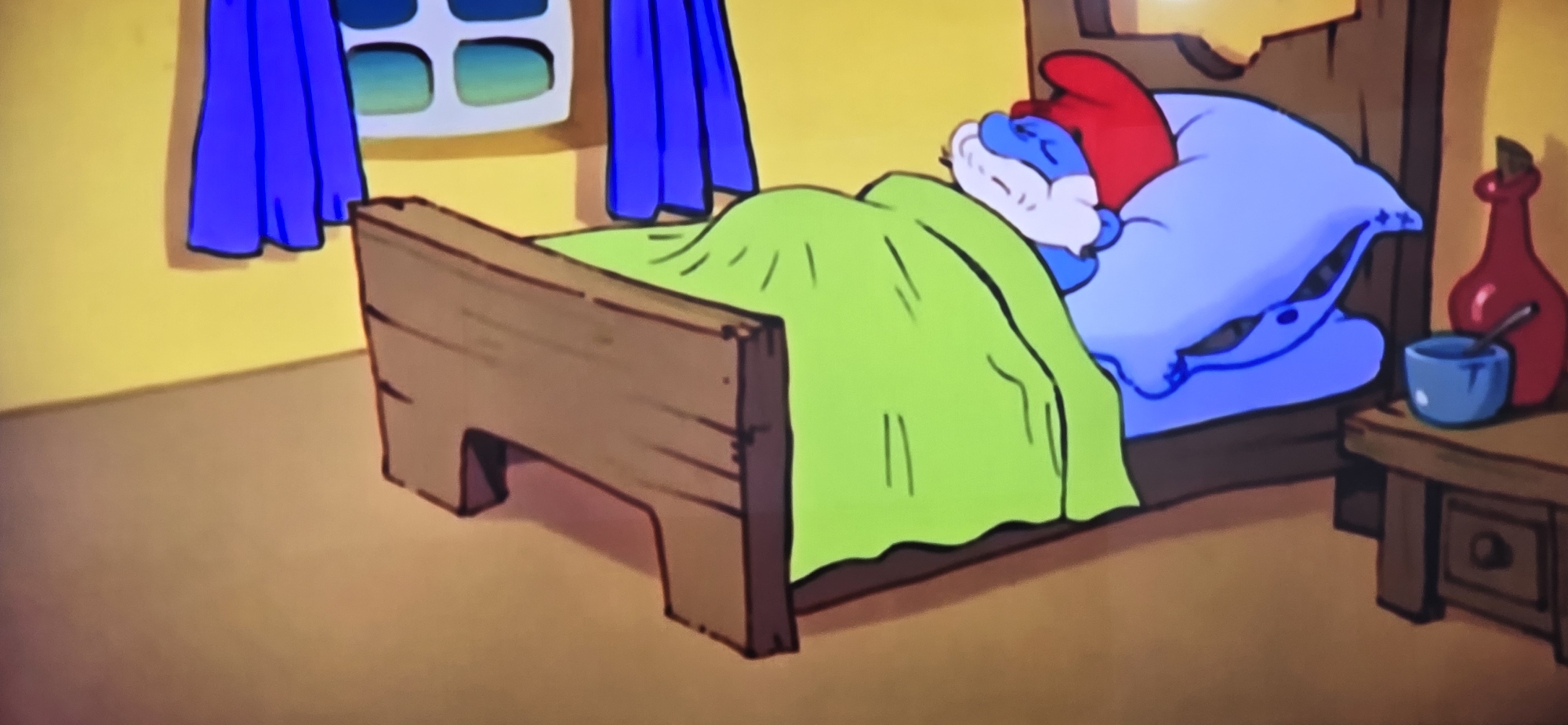 The image depicts a cartoon scene of a Smurf sleeping in a wooden bed. He is covered with a green blanket and has a red hat on. He is sleeping on a blue pillow. To the right of the bed is a small wooden table with a blue cup and a red bottle. In the background is a yellow wall with a window covered by blue curtains. There is no evidence to suggest this image is located in Sofia, Bulgaria.