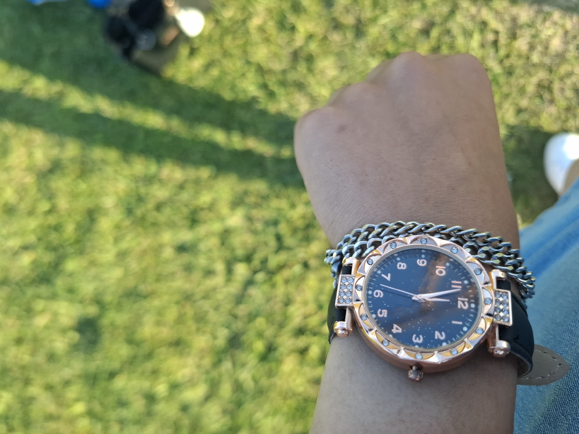 A person's wrist is visible, adorned with a wristwatch and chain bracelets, set against a blurred background of green grass. The watch has a dark face with gold numbers, a gold casing embellished with crystals, and a black leather strap. A silver-colored chain bracelet is layered over the watch. The person is wearing jeans. Part of a white shoe is visible in the right corner. The background is a field of green grass with a blurry collection of objects visible in the top-left. The location is unspecified and cannot be determined as Krapets, Bulgaria, from the given image.