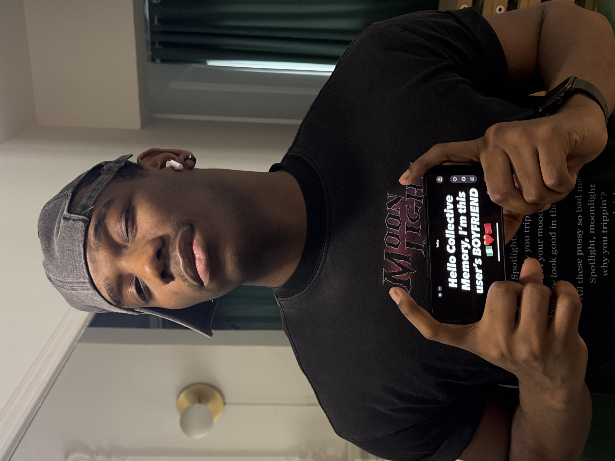 A male individual with dark complexion and short hair is depicted from the chest up, positioned centrally against an indoor background. He is wearing a grey baseball cap backward, a black short-sleeved t-shirt with a "MOON HIGH" graphic, a white wireless earbud in his right ear, and a black ring earring in the same ear. A black smartwatch is visible on his left wrist. He holds a black smartphone horizontally with both hands at chest level. The phone screen displays white text on a black background, which reads: "Hello Collective Memory, I'm this user's BOYFRIEND," followed by the Nigerian flag and the Chinese flag. His expression is direct and unsmiling. The background consists of a light-colored wall with a brass and frosted glass wall sconce on the left, and a partial view of dark green curtains and stacked light-brown objects on the right. A white decorative molding is visible at the top left. This scene is reported to be located in the city of Liangjiang, China.