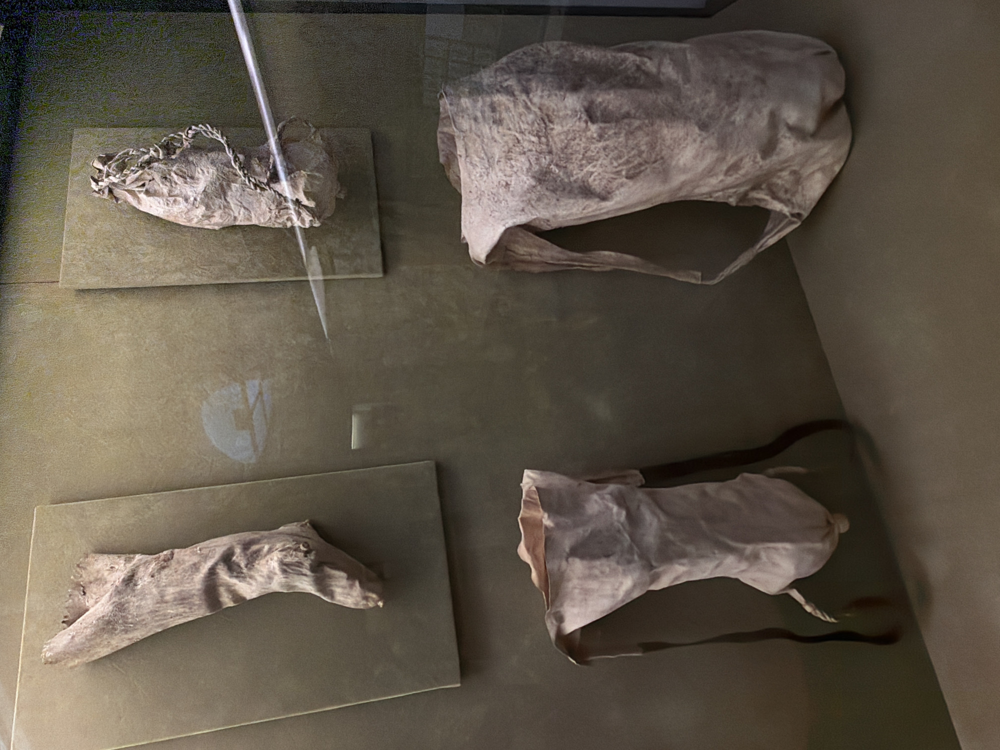 This media file captures a museum display featuring ancient artifacts. The scene is set within a dimly lit exhibition space, likely a museum, with muted green walls serving as the backdrop. The artifacts are presented on rectangular pedestals or stands, some of which appear to be made of glass or polished stone.
The subjects are several aged, preserved objects. In the top left, a pale, cloth-like object with a braided cord or strap across its opening is displayed. To its right, a larger, roughly formed bag or pouch made of similar pale material is shown. In the bottom left, a piece of weathered wood, possibly a tool or a natural object, is laid out. Finally, in the bottom right, another cloth object, resembling a small bag or carrying pouch, is presented with dark straps extending from it. The material of these cloth objects appears brittle and aged, suggesting they are historical artifacts, possibly textiles or leather from an ancient civilization.
There is no discernible activity or event taking place, as the image is a still capture of a static display. The lighting is subdued, creating a somber and historical atmosphere, but does not suggest a specific time of day or weather conditions. The context of Las Palmas De Gran Canaria, Spain, is provided as a location cue, but no specific landmarks or environmental details within the image confirm this. There is no visible text in the image.