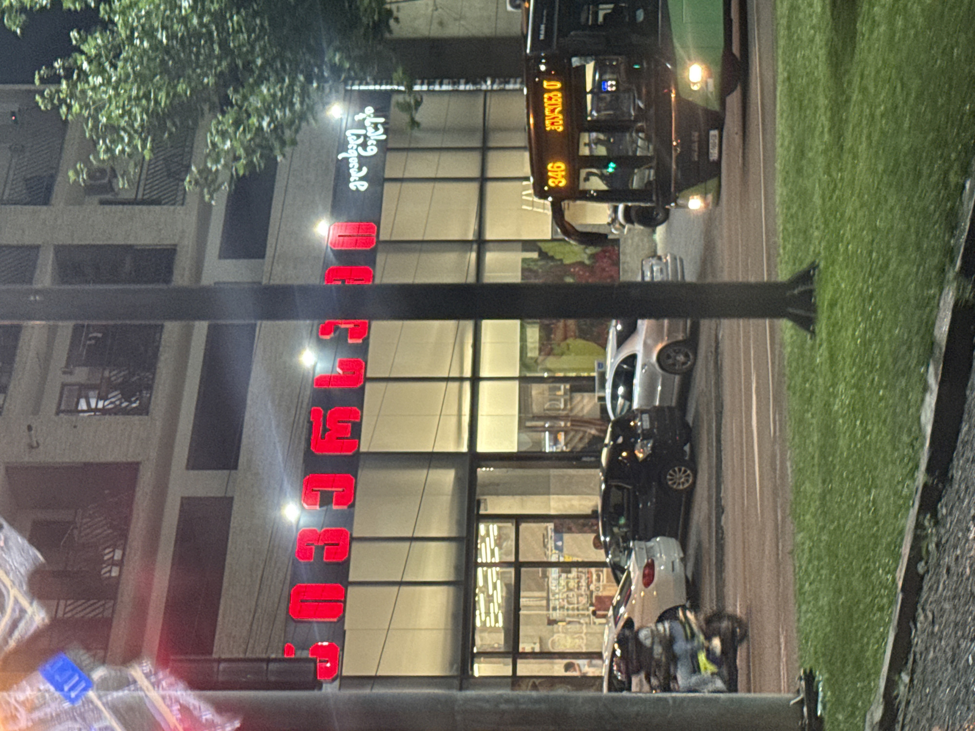 The image shows a street scene at night in Tbilisi, Georgia. A building with a large glass storefront is visible, with bright red Georgian script signage that reads "SPRINT". To the right of the building, a green and black bus with the number "346" illuminated on its front is driving on the street. Several cars are parked or driving on the road, and a person is riding a scooter in the foreground on the left. A tree with green foliage is situated on the right side of the building. The street is lit by the building's interior lights and streetlights, creating a nighttime urban ambiance.