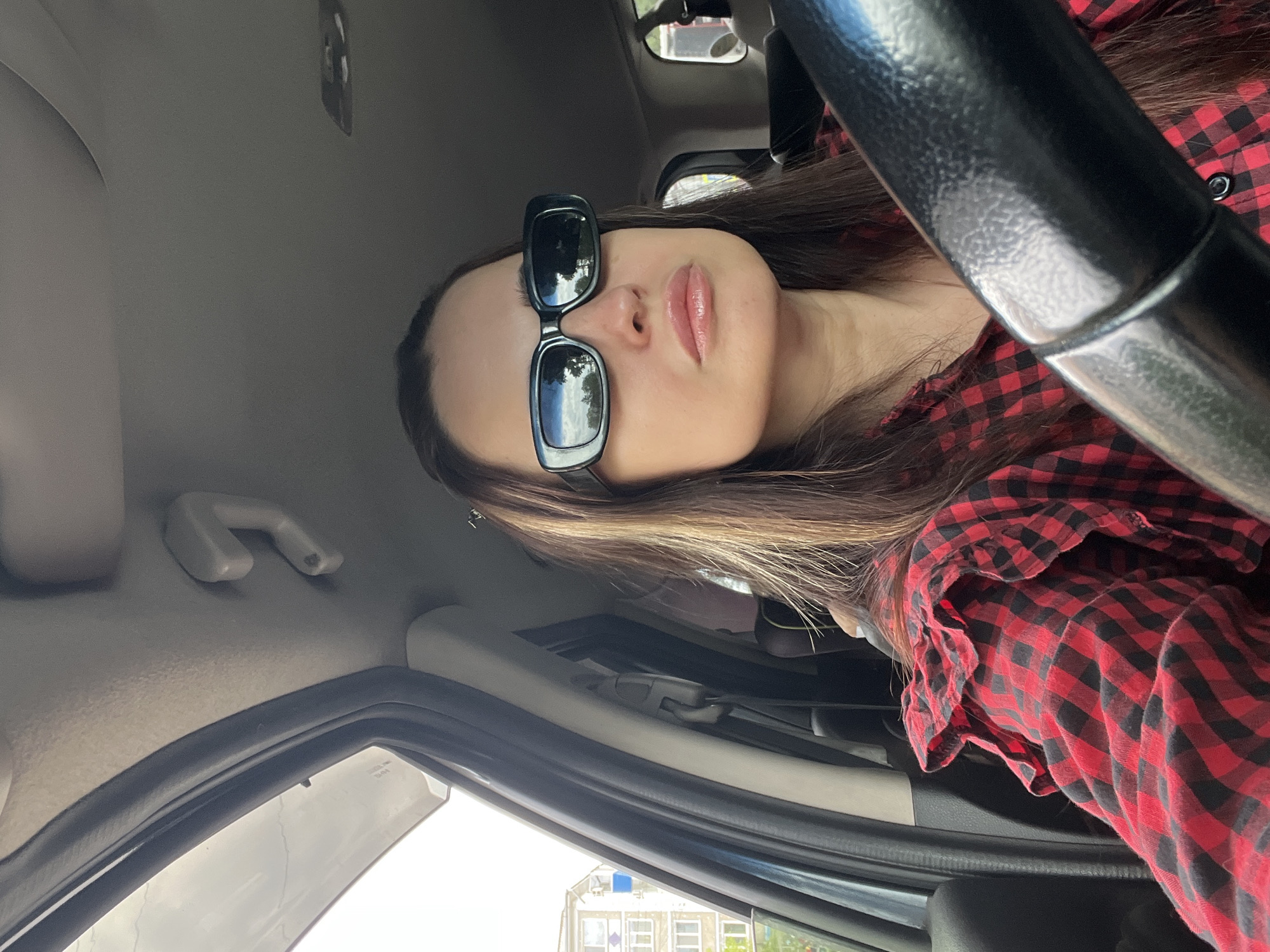 A woman with long brown hair, wearing black-framed sunglasses and a red and black checkered shirt, is seated in the driver's seat of a car. The car's interior is visible, including the steering wheel, dashboard, and window. Outside the car window, a building is visible, suggesting the location is an urban environment. The specific location in the city is unknown.