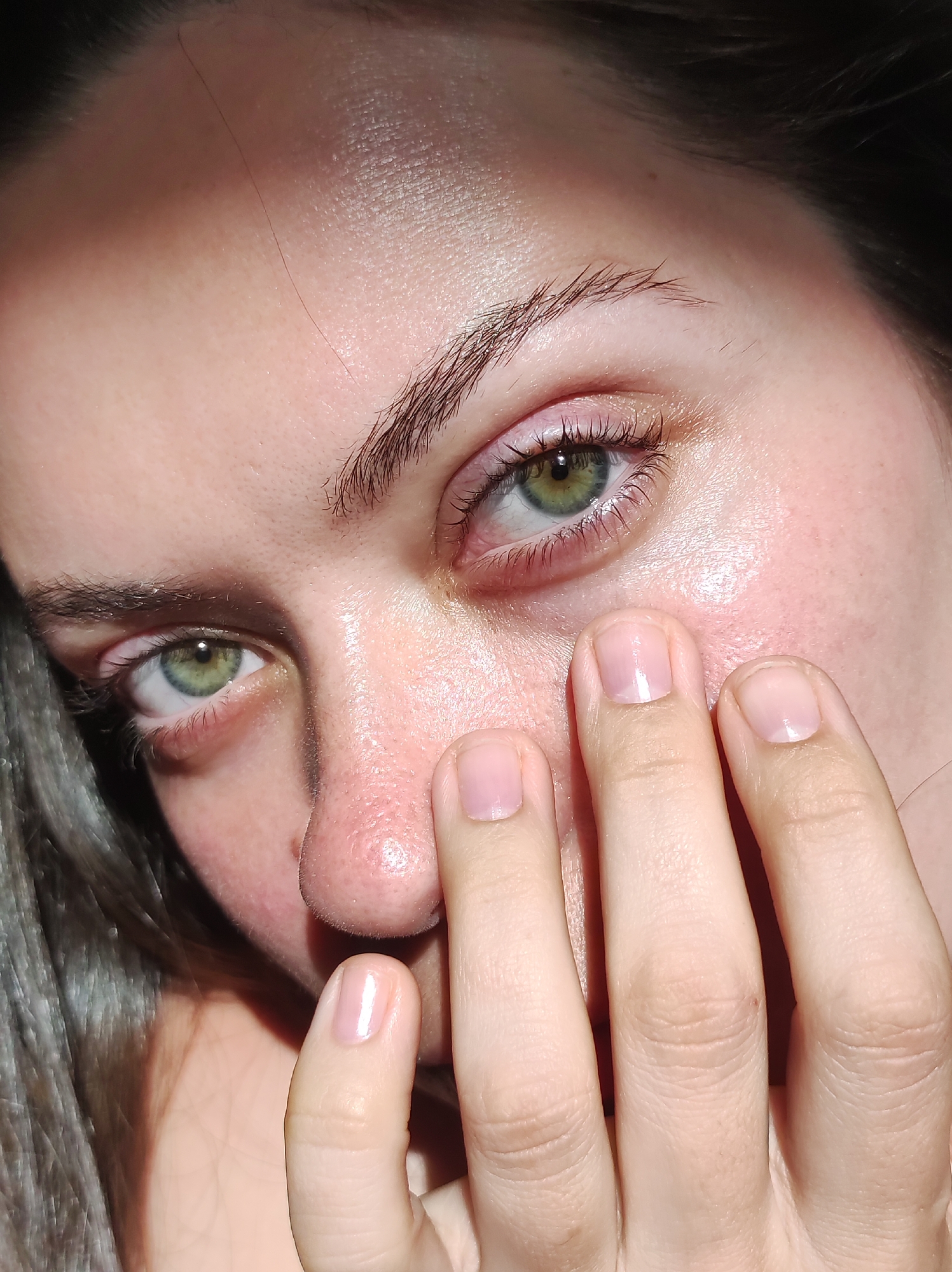 A close-up photograph captures a portion of a person's face. The individual has green eyes, light skin, and light brown eyebrows. A hand is partially covering the face, with fingers touching the cheek and nose. The background is indistinct. The photograph was taken indoors. The location in the city is Unknown, Unknown.