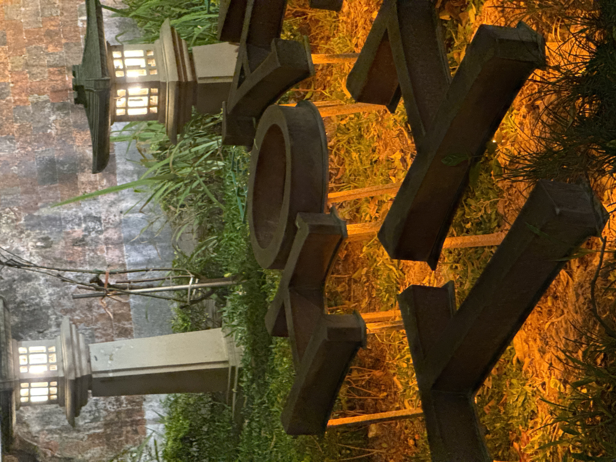 The image shows a close-up of large, dark metal letters spelling out "Y O K Y". The letters are positioned vertically and angled, suggesting they might be part of a sculpture or signage. The metal appears weathered and aged. Behind the letters, there is dense green foliage interspersed with dry, orange-toned vegetation, illuminated by a warm, artificial light source, possibly from landscape lighting. The lighting creates a dramatic and somewhat moody atmosphere.
To the left of the sculpture, two outdoor lanterns with illuminated panes are visible. These lanterns have a modern, geometric design and are placed on either side of a textured, brick-like wall. The wall has variations in color, ranging from muted reds to grays, and appears somewhat aged. The presence of the lanterns and the brick wall suggests an outdoor setting, possibly a garden, walkway, or entrance. The foliage, along with the overall aesthetic, could indicate a location in Southeast Asia, with the mention of Hanoi, Vietnam, providing specific context. The time of day appears to be evening or night due to the artificial lighting and the darkness of the background. No people are visible in the image, and the focus is entirely on the sculptural letters, lanterns, and the surrounding environment.