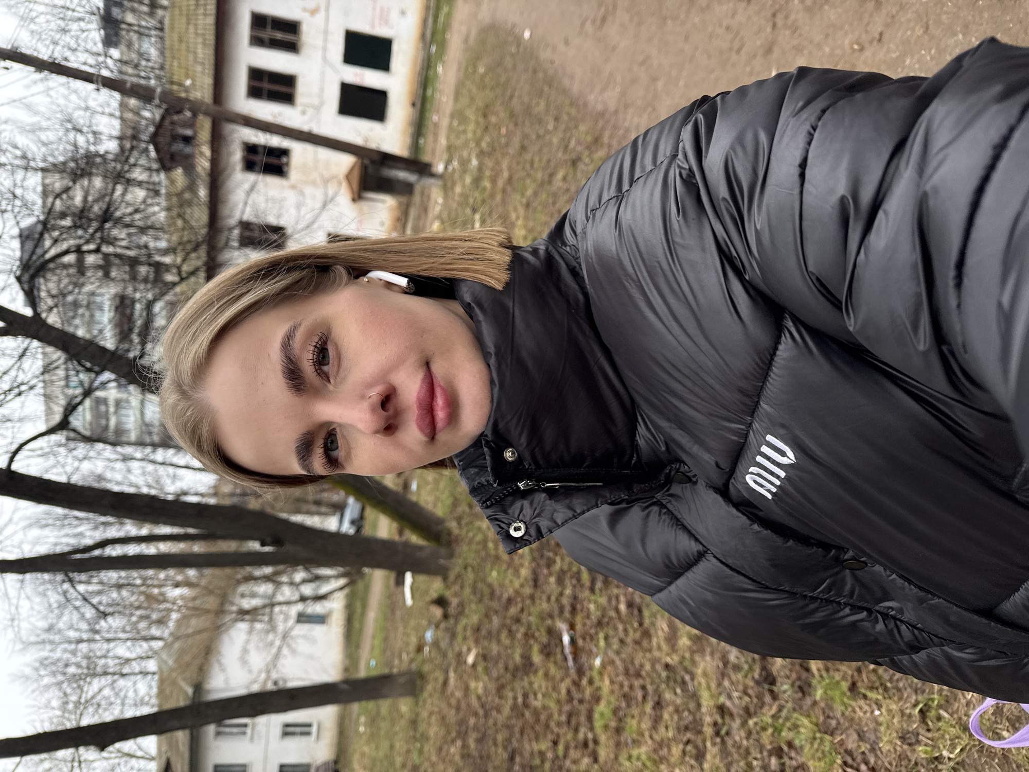 A young woman wearing a black puffer jacket with "MIU" embroidered on the right breast is looking at the camera. She is wearing an earbud in her right ear and has a nose ring. The background shows trees with bare branches, a patch of ground with dried leaves, and a white building with dark windows. The photograph appears to be taken in an urban environment, specifically the city of Chaikovsky, Russia.