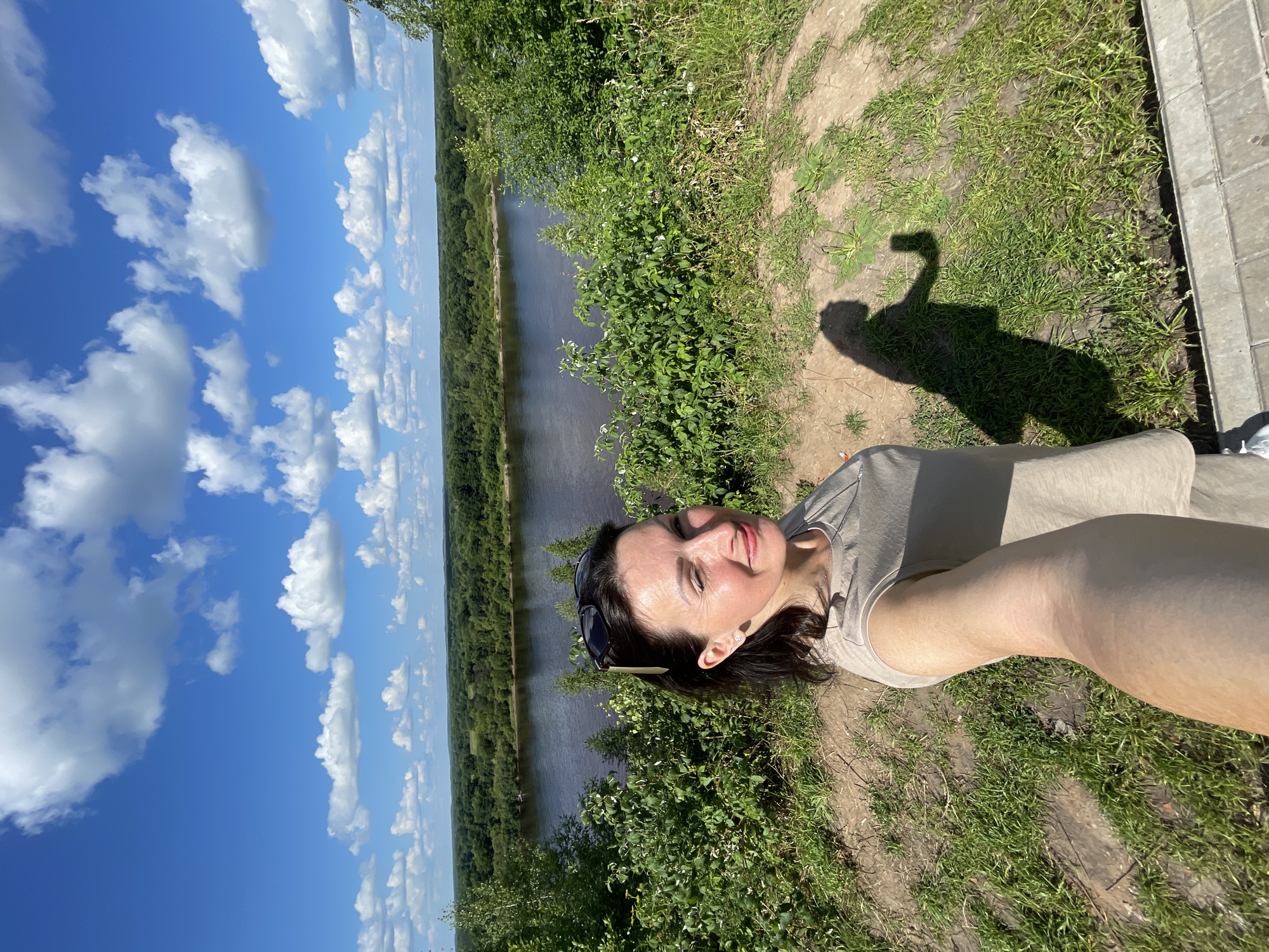 A woman is taking a selfie on a sunny day with a river and forest in the background. She is standing on a grassy hill overlooking a wide river. The sky is blue with white clouds. The woman has dark hair, is wearing a light-colored t-shirt, and has sunglasses on top of her head. Her shadow is visible on the ground. The location is the picturesque embankment of the Cheptsa River in Kirovo-Chepetsk, Russia.