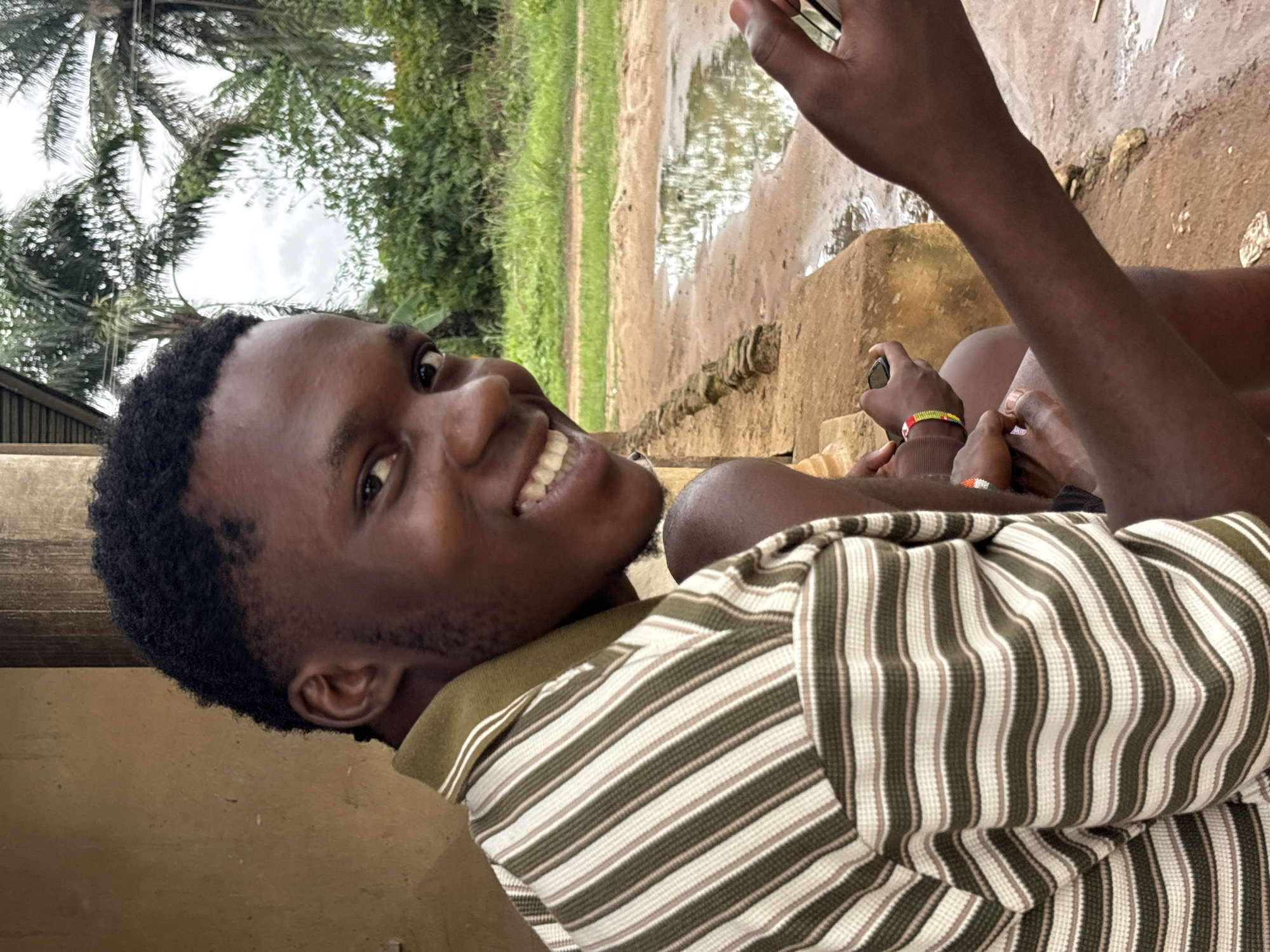 A young, dark-skinned man with short hair and a stubbly beard smiles at the camera. He is wearing a striped shirt and appears to be sitting. In the background is a muddy, rural environment with trees and a puddle. The location is likely Ikot Ekpene, Nigeria, based on the prompt. In the upper right corner, another person is visible, holding a phone in one hand and wearing bracelets on the other.