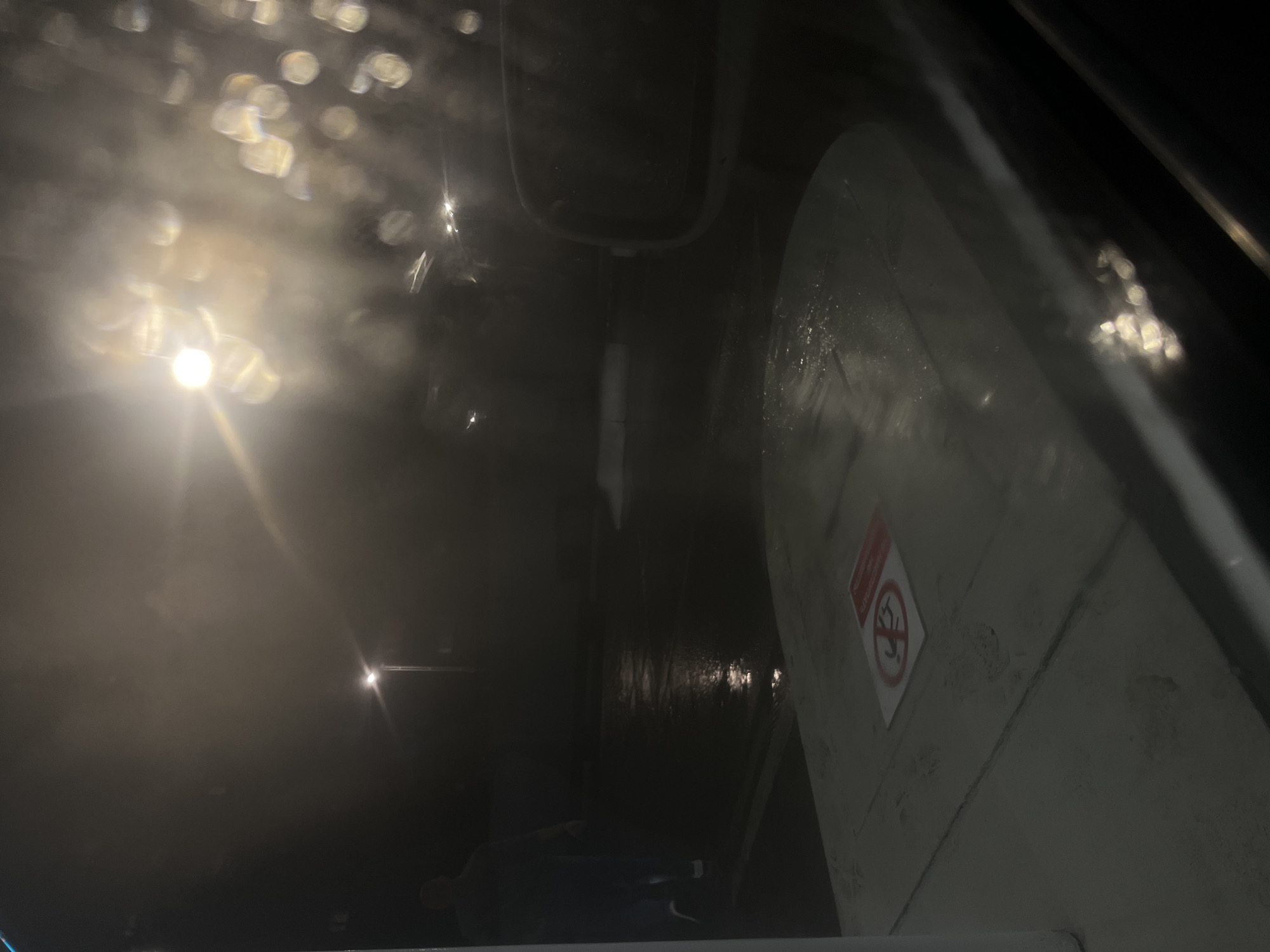 In Belogorsk, Russia, at night, an outdoor scene unfolds through a wet windshield. A person, silhouetted and blurred, walks away from the camera on a wet, reflective asphalt surface. Streetlights illuminate the foggy, dark environment, casting a hazy glow and creating specular highlights on the wet ground. A car side mirror is visible in the right foreground. In the lower left, a concrete surface with a "no smoking" sign is partially visible. The overall impression is one of a dark, damp, and somewhat obscured urban setting.