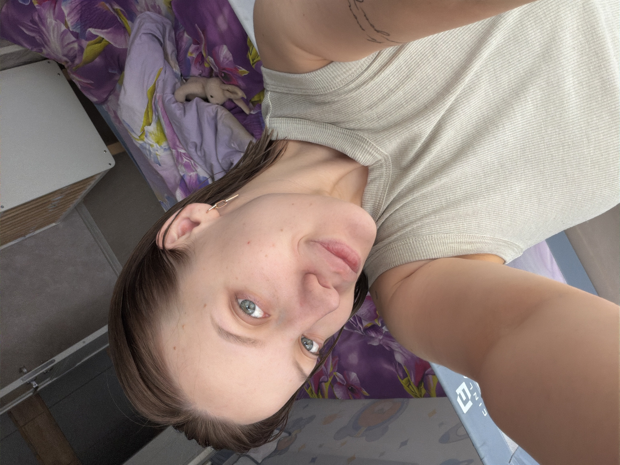 An image captures a young adult female in what appears to be a bedroom setting, likely taken as a selfie from an overhead, downward-looking angle. She is lying down or reclining, looking directly at the camera with a slight, calm smile. Her light-colored eyes (blue/grey) are open, and she has light brown hair, fair skin with some small freckles, and a visible tattoo on her upper left arm. She wears a light-colored, possibly beige or off-white, ribbed sleeveless top and a gold-colored earring in her left ear.
The background shows a bed with purple and green floral patterned bedding. A small, light-colored stuffed animal, resembling a rabbit, is partially visible on the bedspread. To the left, a simple white bedside table or surface is present. In the lower right corner, a portion of a blue and white patterned sheet or blanket is visible, featuring partial text that appears to be "LIU JIN" or similar characters. The overall lighting suggests it is daytime. The location context provided is Reshetnikovo, Russia.