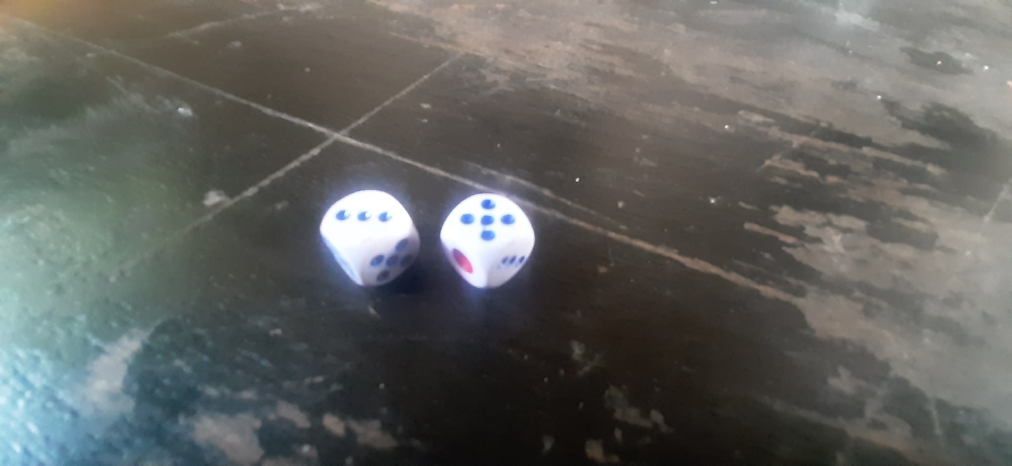The image shows two white dice with blue pips lying on a dark, textured surface. The surface appears to be wood, possibly a table, with visible grain and some wear marks. The dice are positioned close together in the lower-left quadrant of the image.
The die on the left shows a face with five pips arranged in a cross pattern, and another face with four pips arranged in a square. The die on the right shows a face with five pips, and a partial view of a face with three pips. One of the dice also has a red pip, suggesting it might be a special type of die or there's a red marking on it. The lighting suggests it's daytime, but there are no specific cues about the time of day. The background is out of focus, making it difficult to determine the exact location, but the context provided suggests Yessentukskaya, Russia. No people are visible in the image, nor is there any text. The activity taking place is simply the dice resting on the surface.