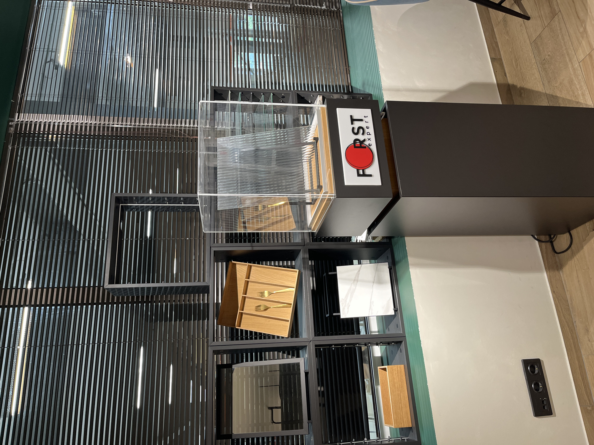The image shows an interior display of kitchen cabinet organizers and accessories. The scene is dominated by a modern, dark-colored shelving unit with various organizers displayed within. These include wooden cutlery dividers, a marble tile sample, a mirrored panel, and a taller wooden storage container. At the top of the unit, there is a display showcasing ribbed glass or acrylic sheets, with a branded sign "FIRST expert" next to it.
The backdrop features a slatted design, possibly a window covering or decorative wall. The background also reveals sections of a light green wall, a beige wall, and a wooden floor. The environment is indoors and well-lit. The image does not feature any people or interactions.