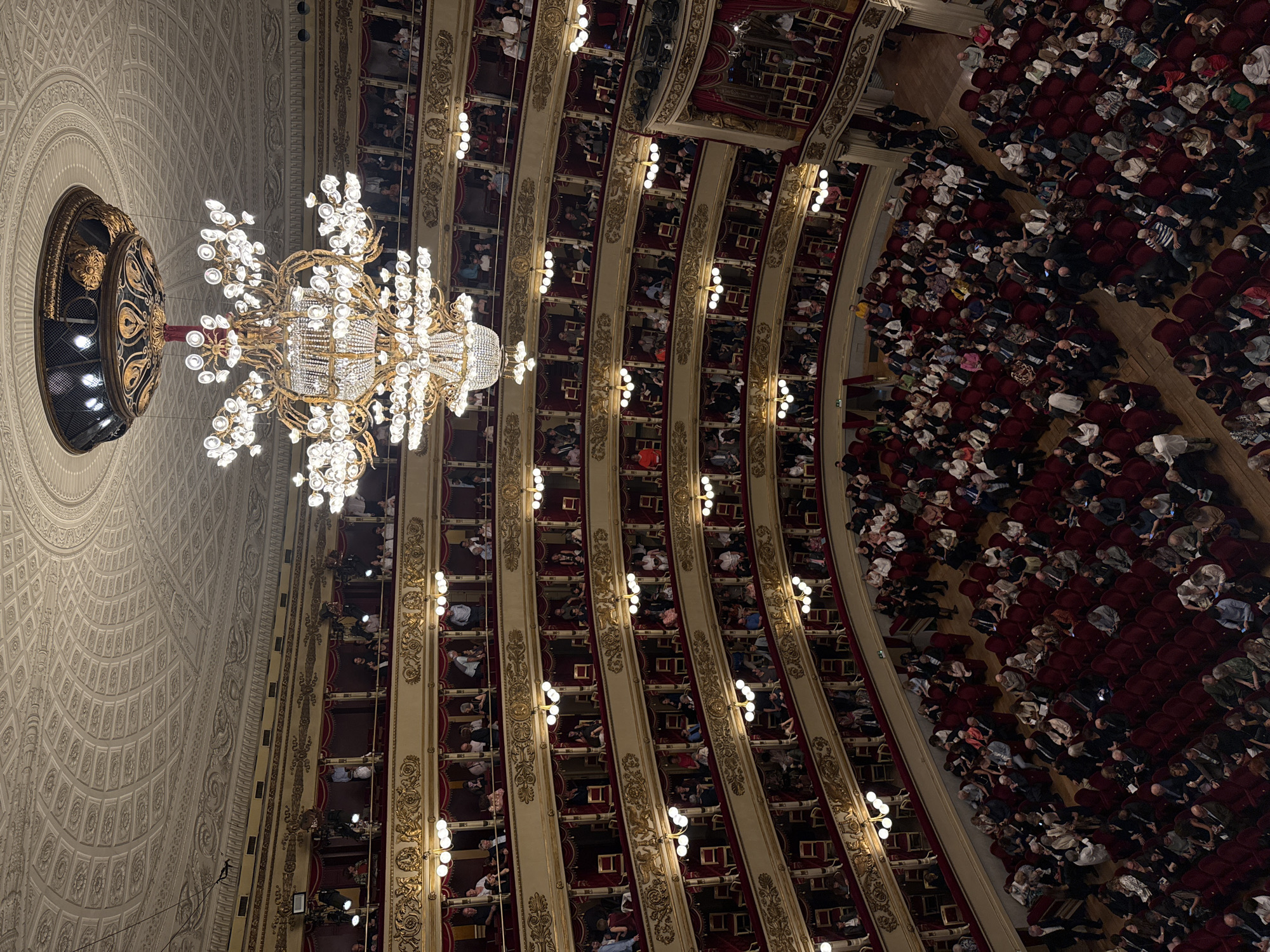 The image depicts the interior of a grand opera house, specifically the Teatro alla Scala in Milan, Italy. The perspective is from above, looking down into the theater. A large, ornate chandelier with multiple lights hangs prominently in the center of the frame, illuminating the opulent interior. The theater features tiered seating boxes with red velvet upholstery and gilded trim, characteristic of historical European opera houses. In the boxes and on the main floor, a crowd of people is seated, dressed in formal attire, suggesting they are attending a performance. The ceiling is elaborately decorated with intricate moldings and patterns. The overall impression is one of grandeur, elegance, and a cultural event in progress.