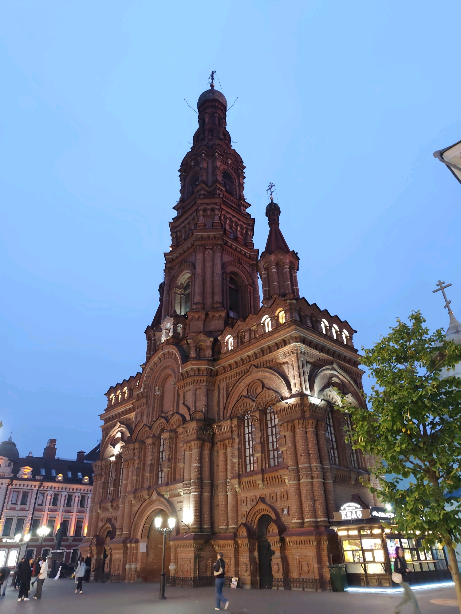 This is a low-angle, full-shot photograph of the exterior of the Peter and Paul Cathedral in Kazan, Russia, captured during twilight. The large, ornate brick cathedral dominates the center of the frame, its intricate details illuminated by interior lighting, highlighting its tiered structure and multiple spires topped with crosses. To the left of the cathedral, several people are walking on a paved plaza, some conversing and others looking at their phones. The plaza is lined with streetlights and bordering buildings with illuminated windows. To the right of the cathedral, a lush green tree partially obscures a silver-domed structure topped with a cross. The sky is a deep, clear blue, indicative of the evening hour. The overall scene depicts a lively urban environment with the majestic cathedral as the central focal point.