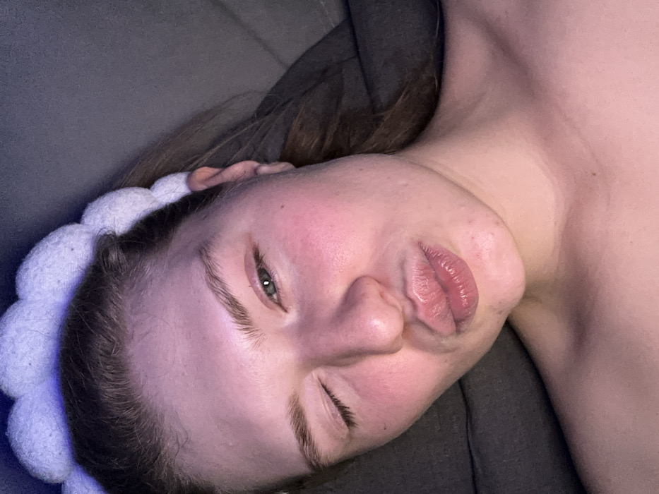 A pale-skinned person, likely female, reclines on a gray surface, possibly a couch. They wear a white, textured headband. Their hair is brown and partially obscured. The person’s face is angled toward the camera, with one eye partially closed in a wink. Their lips are pursed into a kiss, creating wrinkles around the mouth. The skin on their face appears slightly flushed. The overall image is dimly lit, creating shadows and highlights across the face and neck.