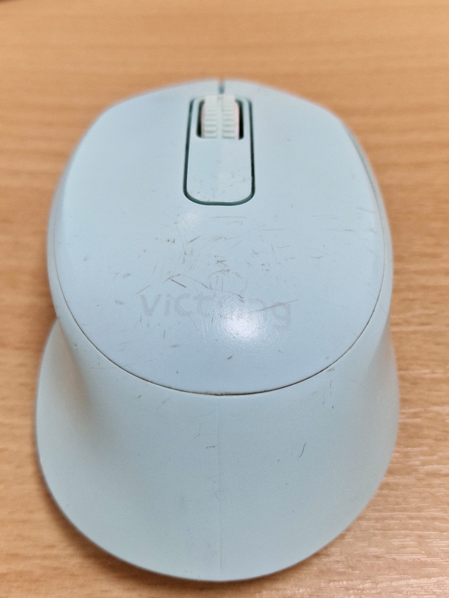 A light blue computer mouse with a gray scroll wheel is positioned on a light brown wooden surface. The mouse has faint scratches and a translucent logo that reads "VICTORY". The scroll wheel is recessed and has a textured surface.