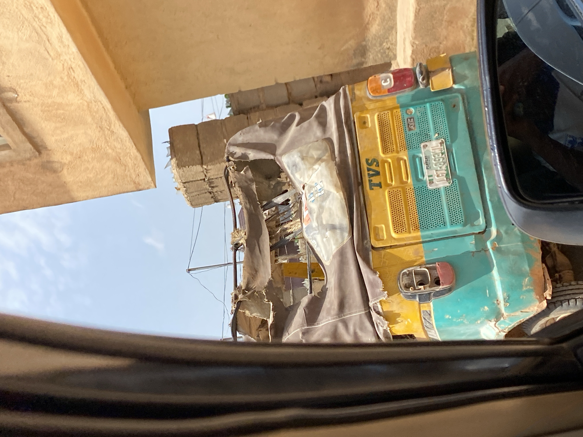 In Gura Top, Nigeria, a damaged yellow and teal auto-rickshaw, bearing the "TVS" logo and license plate "LGT-893NL", is parked outdoors. The fabric covering the auto-rickshaw's roof is ripped and torn, exposing its internal structure. Adjacent to the auto-rickshaw, a tan building with a window and a ledge is visible. Stacked bricks are also present in the vicinity, and power lines are strung across the sky. The scene appears to be captured from inside another vehicle, as evidenced by a side-view mirror reflecting a person and a steering wheel. The overall environment suggests a dusty, possibly arid, urban setting.