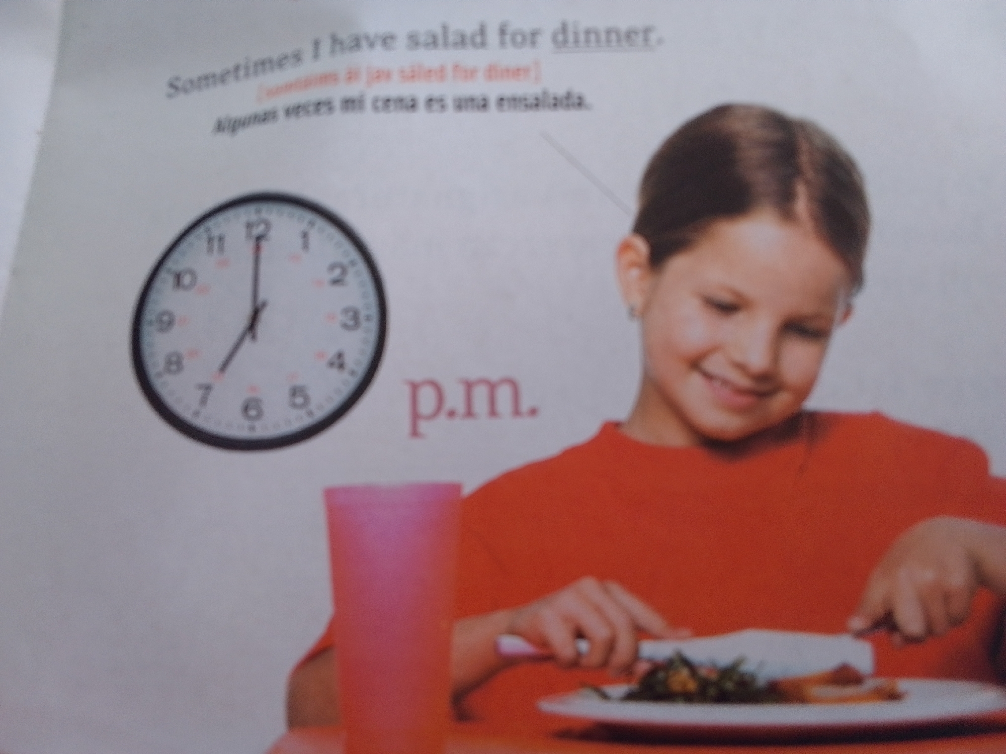 This media file features a young girl eating salad for dinner. She is wearing a bright orange shirt and is seated at a table with a pink cup and a plate of food. A clock on the wall indicates the time is around 6:45 p.m. The background text, in English and Spanish, translates to "Sometimes I have salad for dinner." The overall setting appears to be indoors, possibly a home or a classroom, given the educational nature of the text. The girl has a happy expression, suggesting enjoyment of her meal. There are no clear indicators of the weather or specific location beyond the provided context of La Tagua, Venezuela.