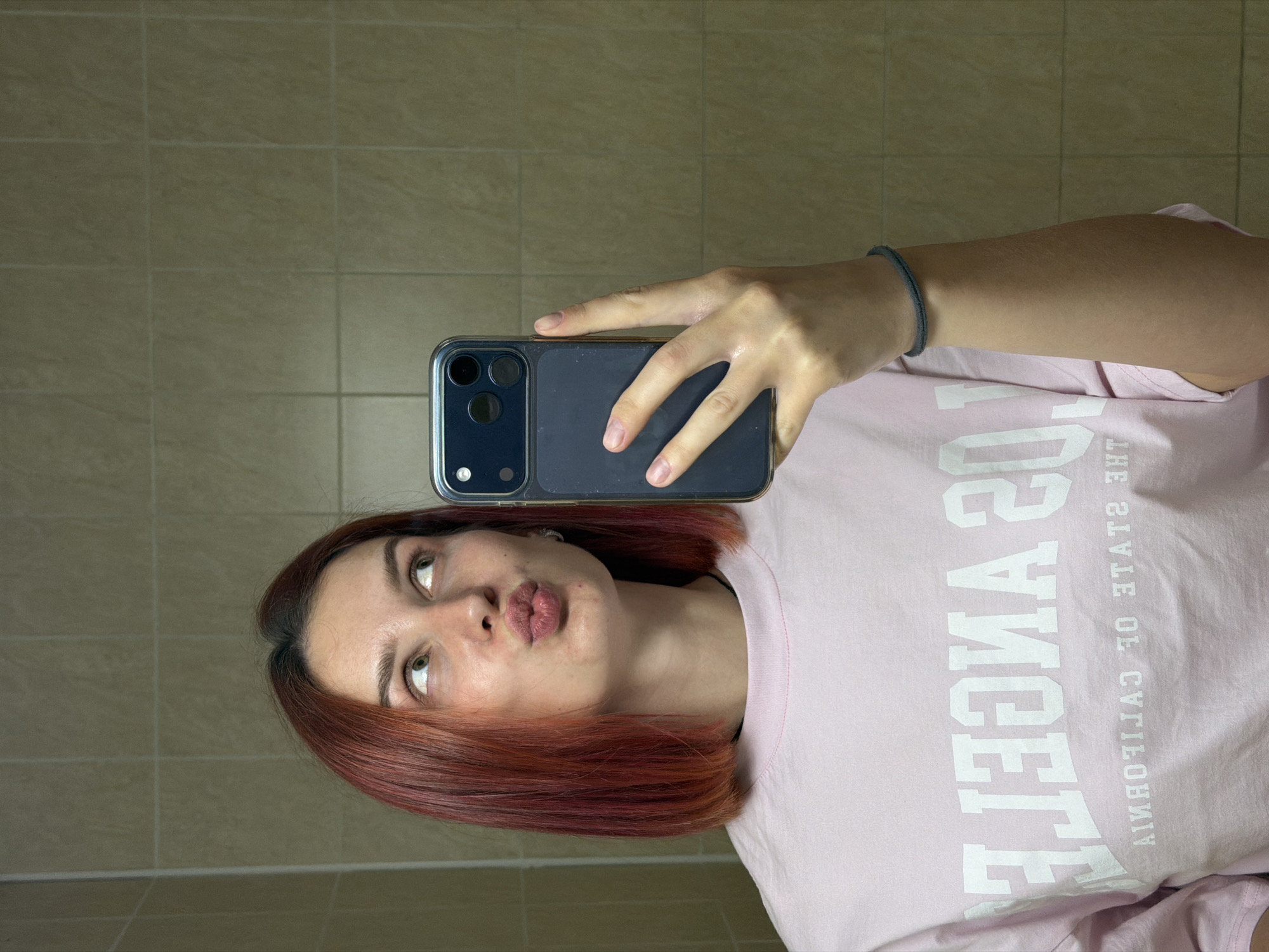 A person is taking a selfie in what appears to be a bathroom. The individual has auburn, shoulder-length hair and is wearing a light pink t-shirt with "Los Angeles, the state of California" printed on it. They are holding a dark-colored smartphone with a triple-lens camera in their right hand, positioning it to capture their face. The person has a slight "duck face" expression. The background consists of tiled walls, indicative of a typical bathroom setting. There is no indication of the location being Naberezhnye Chelny, Russia or any other location.