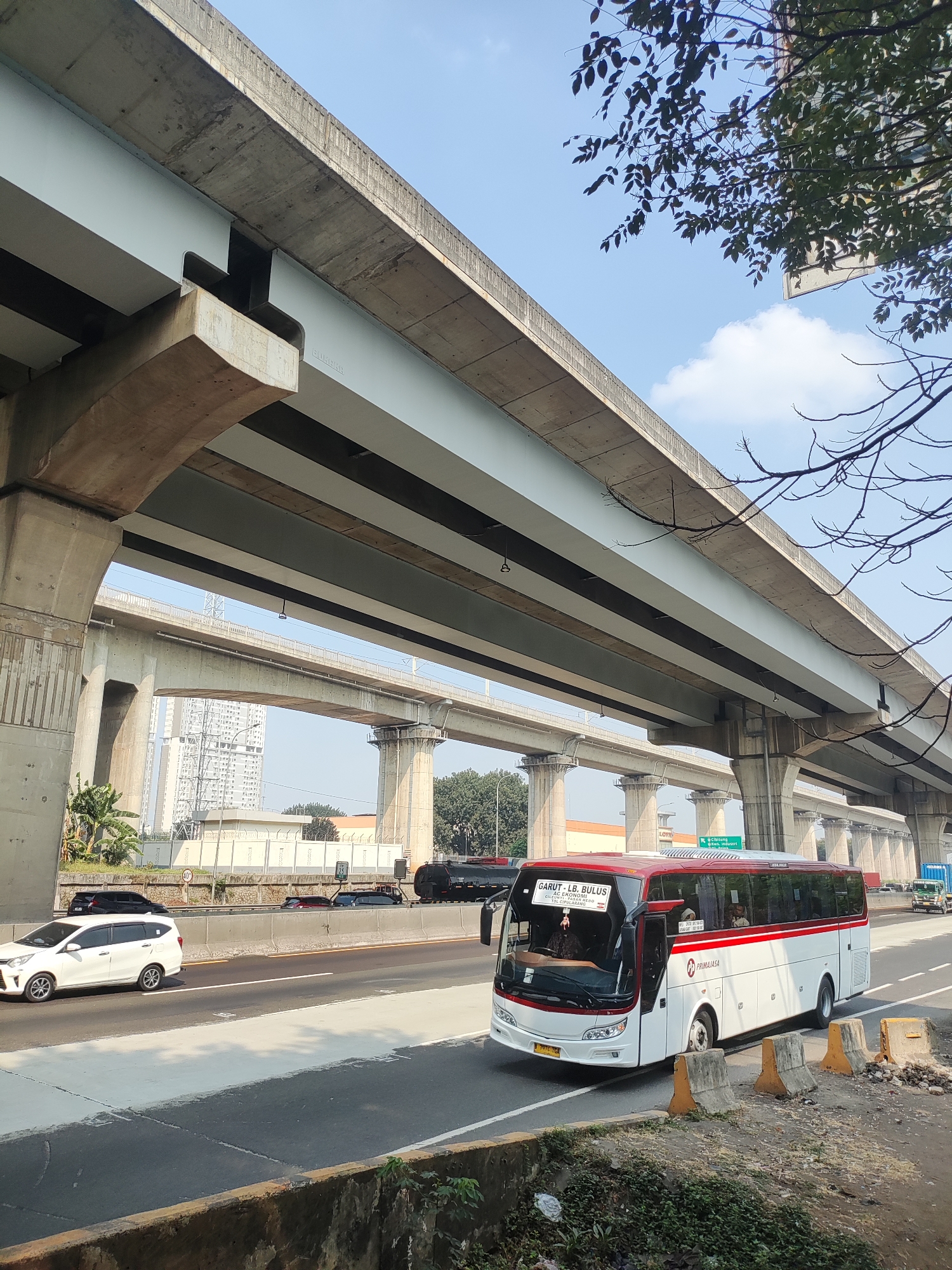 A white and red bus, with the text "GARUT - LB. BULUS" on the front, travels on an elevated highway in an urban environment. The bus has a "PRIMAJASA" logo on its side. The highway is supported by large concrete pillars, and other vehicles, including a white sedan and a black truck, are visible on the road. In the background, tall buildings and trees can be seen, along with a clear blue sky.