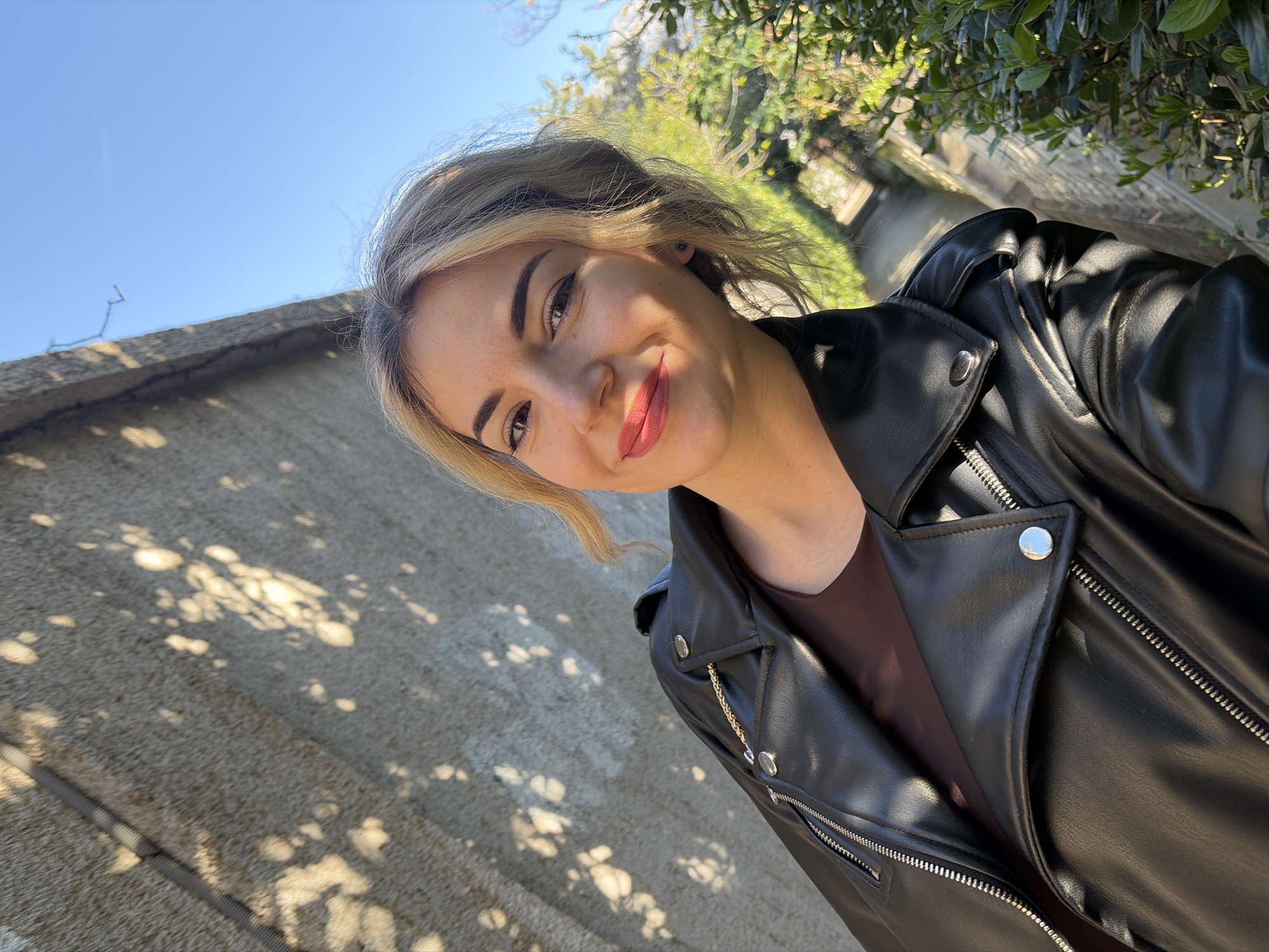 An outdoor self-portrait features a female subject in Budva, Montenegro. The woman, appearing to be in her late 20s to early 30s, is centered, looking directly at the camera with a subtle smile. She has light-colored hair with darker roots, defined eyebrows, and lips colored with reddish-pink lipstick. She wears a black faux leather jacket with silver zippers and snap buttons, over a dark brown or maroon top. A golden chain accessory is visible on the left shoulder of her jacket. The background on the left consists of a textured, light grey concrete wall, displaying dappled patterns of sunlight and leaf shadows. Above the wall and to the upper right is a clear blue sky. To the mid-right and lower-right, dense green foliage, consistent with bushes or hedges, borders a light-colored, sunlit pathway extending into the background. The scene is illuminated by strong, direct sunlight, creating distinct shadows.