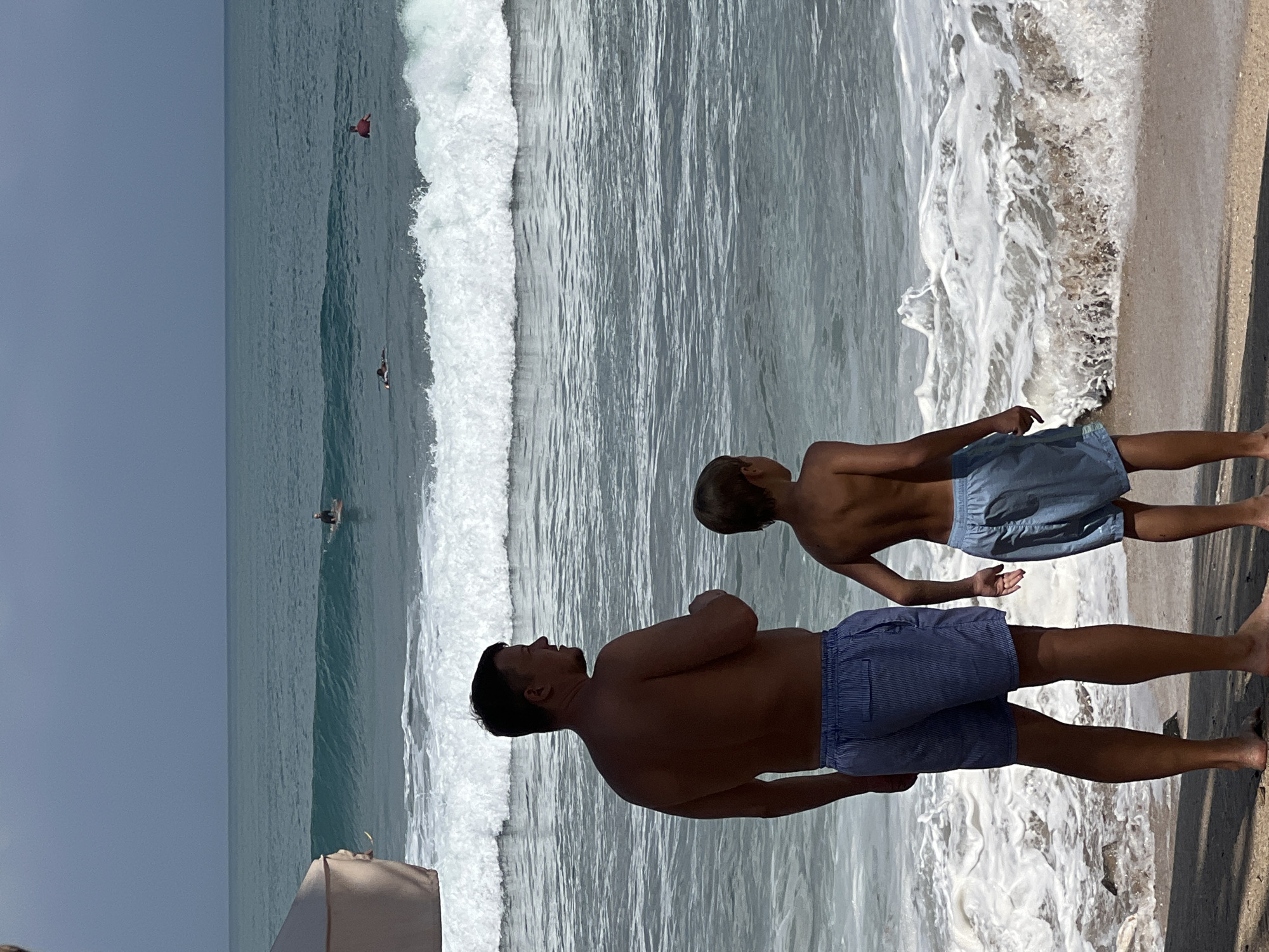 In Kuta, Bali, Indonesia, two individuals, an adult male and a child, stand on the beach facing the ocean. The adult male, with dark hair, wears blue striped shorts, and the child, also with dark hair, wears blue shorts. Both are shirtless. The ocean is active with moderate waves and white foam breaking near the shore. Several surfers are visible in the distance, riding the waves. The sky is overcast, and a portion of a light-colored beach umbrella is visible in the upper left corner of the frame. The sand is visible in the foreground, and the water transitions from foamy white near the shore to darker blue further out.