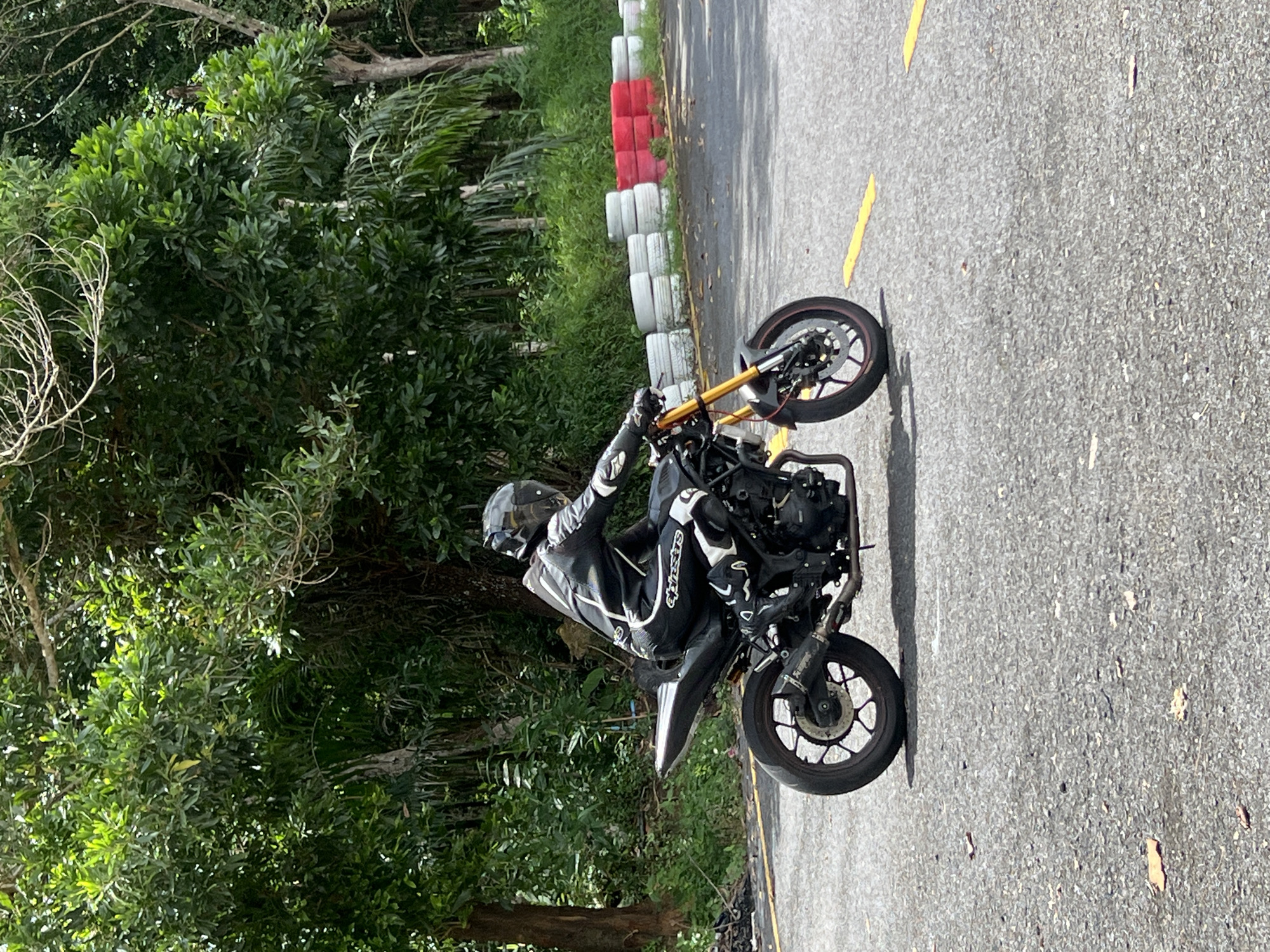A person wearing a black and white motorcycle suit and helmet is riding a black motorcycle with gold-colored front suspension on an asphalt road. The motorcycle has a custom exhaust. The road has yellow lines painted on it. In the background, there is a line of stacked tires, some white and some red, acting as a barrier. Beyond the tires is lush green vegetation, including trees and bushes. The scene appears to be outdoors, possibly on a track or road in a natural environment. The location is identified as Thep Krasatti, Thailand.