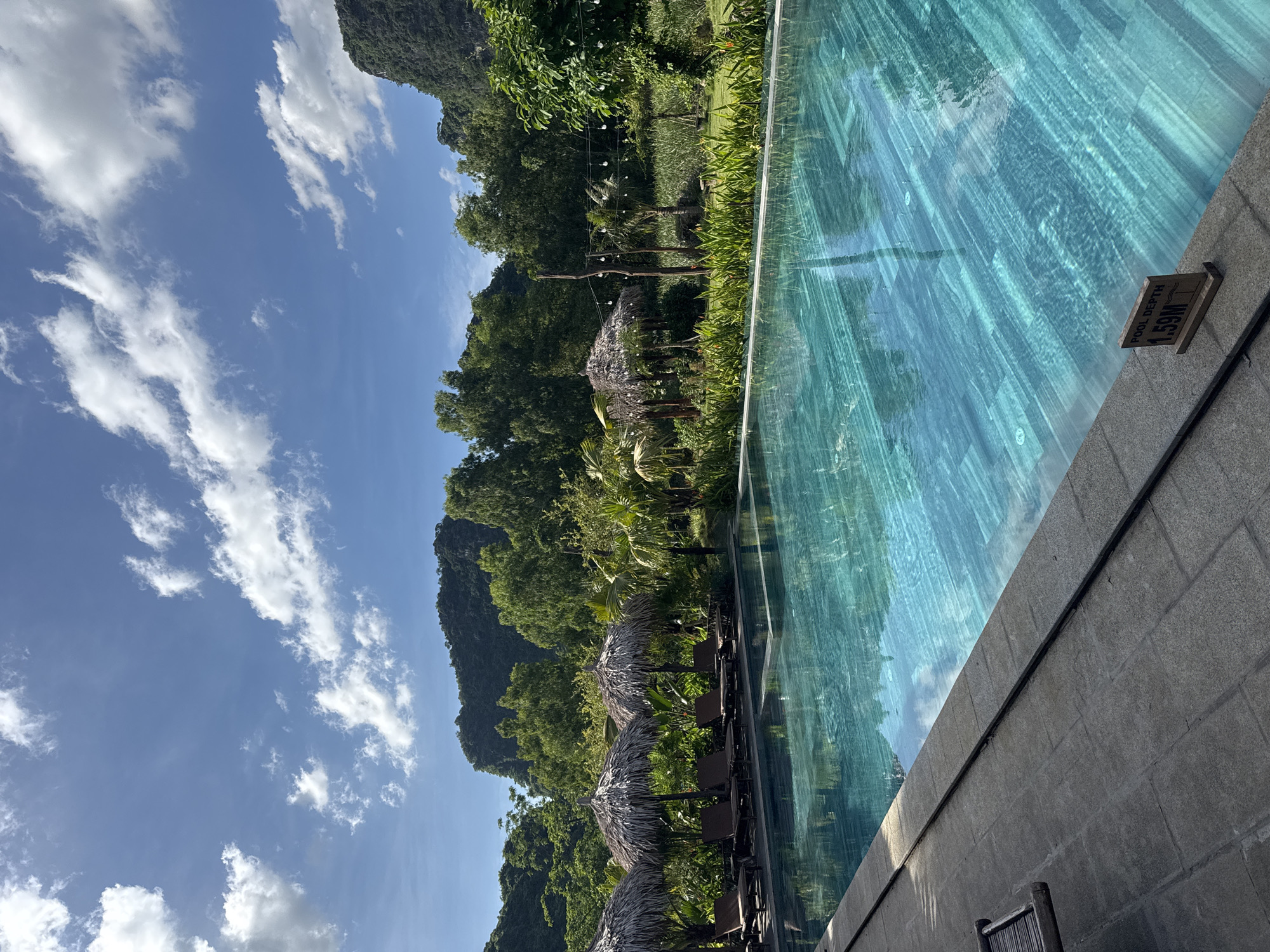 This image captures a serene outdoor swimming pool area in Nam Hoa Lư Ward, Vietnam, under a bright, partially cloudy sky. The pool itself is a striking turquoise, with a textured bottom resembling light-colored wood planks arranged diagonally. The water's surface reflects the sky and surrounding greenery.
Along the edge of the pool, a stone walkway is visible, with a small wooden sign indicating "POOL DEPTH 1.59M". To the left of the pool, several lounge chairs are arranged under thatched-roof umbrellas, suggesting a relaxation area.
The background is dominated by lush, dense green foliage, including various trees and tropical plants. Behind the greenery, majestic limestone karst mountains rise, their slopes covered in dense forest. In the middle ground, a line of string lights is suspended between poles, adding a decorative element to the landscape. The overall impression is one of tranquil tropical resort ambiance.