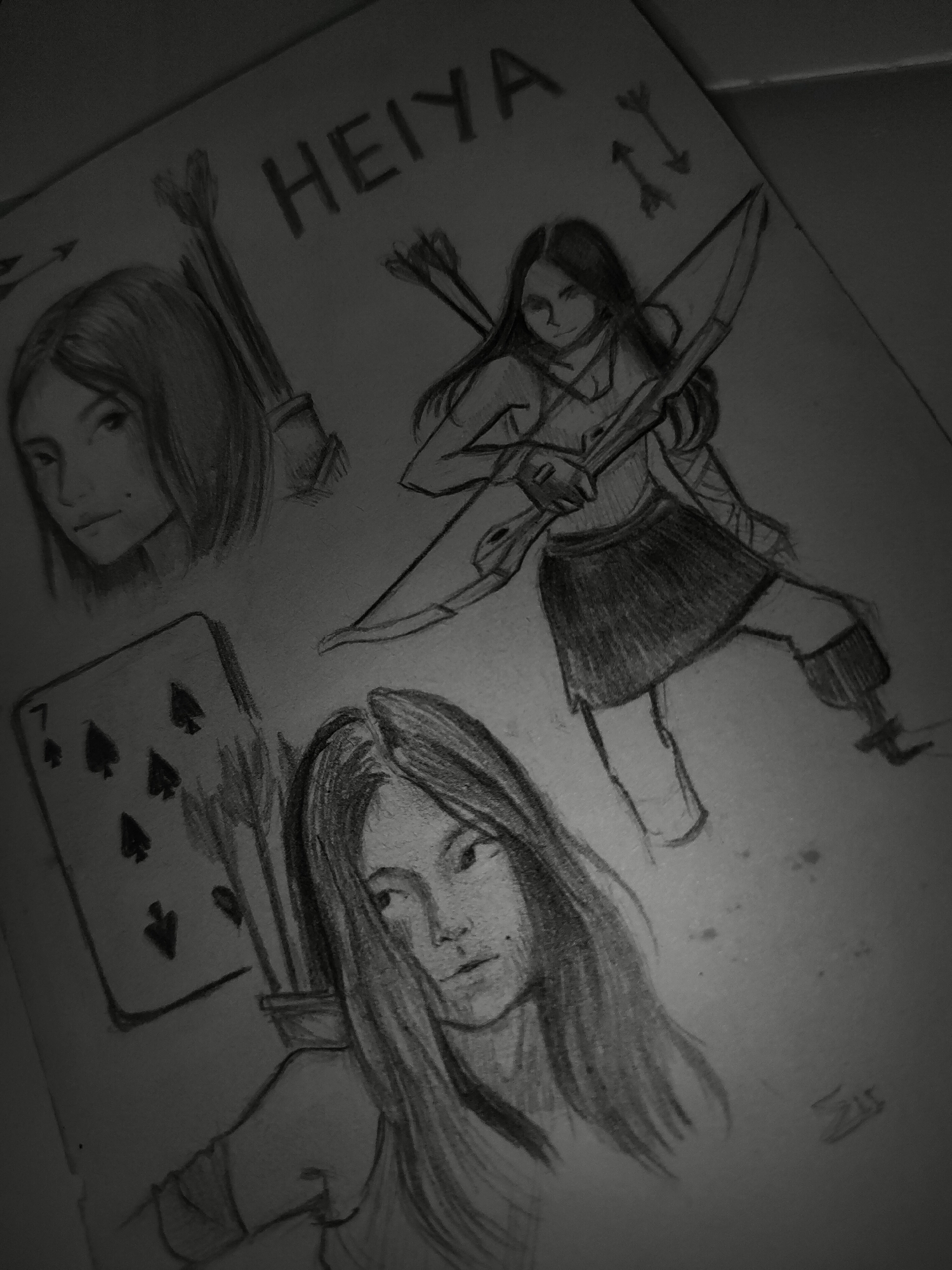 The image is a monochromatic photograph of a pencil sketch on a sheet of paper. The paper is diagonally oriented, with its upper left and lower right corners slightly out of frame. The overall composition is a collection of related drawings.
At the top-center of the paper, the capitalized word "HEIYA" is hand-drawn. Below and to the right of this title, two small, stylized arrows are sketched.
The primary subject is a female character, depicted three times. The largest depiction, centrally located, is a full-body sketch of the character in a dynamic pose, holding a recurve bow with an arrow drawn, ready to fire. She has long, dark hair, wears a sleeveless top, a pleated skirt, and wraps on her lower legs.
To the upper-left of the central figure, a head-and-shoulders portrait of the same character shows her with a neutral expression, looking slightly to the left. A quiver filled with arrows is visible behind her left shoulder.
To the lower-center, another head-and-shoulders portrait shows the character looking to her right, with a more contemplative expression. Again, a quiver of arrows is visible behind her right shoulder.
To the left of this lower portrait, a playing card, specifically the 7 of Spades, is drawn vertically.
In the lower-right corner of the paper, a small, stylized signature or set of initials is present.
The drawings are executed with varying pencil pressures, creating depth and shadow. The background of the photograph is dark and out of focus, indicating a shallow depth of field or low light conditions, and does not provide any identifying information regarding a specific location such as Davao City, Philippines.