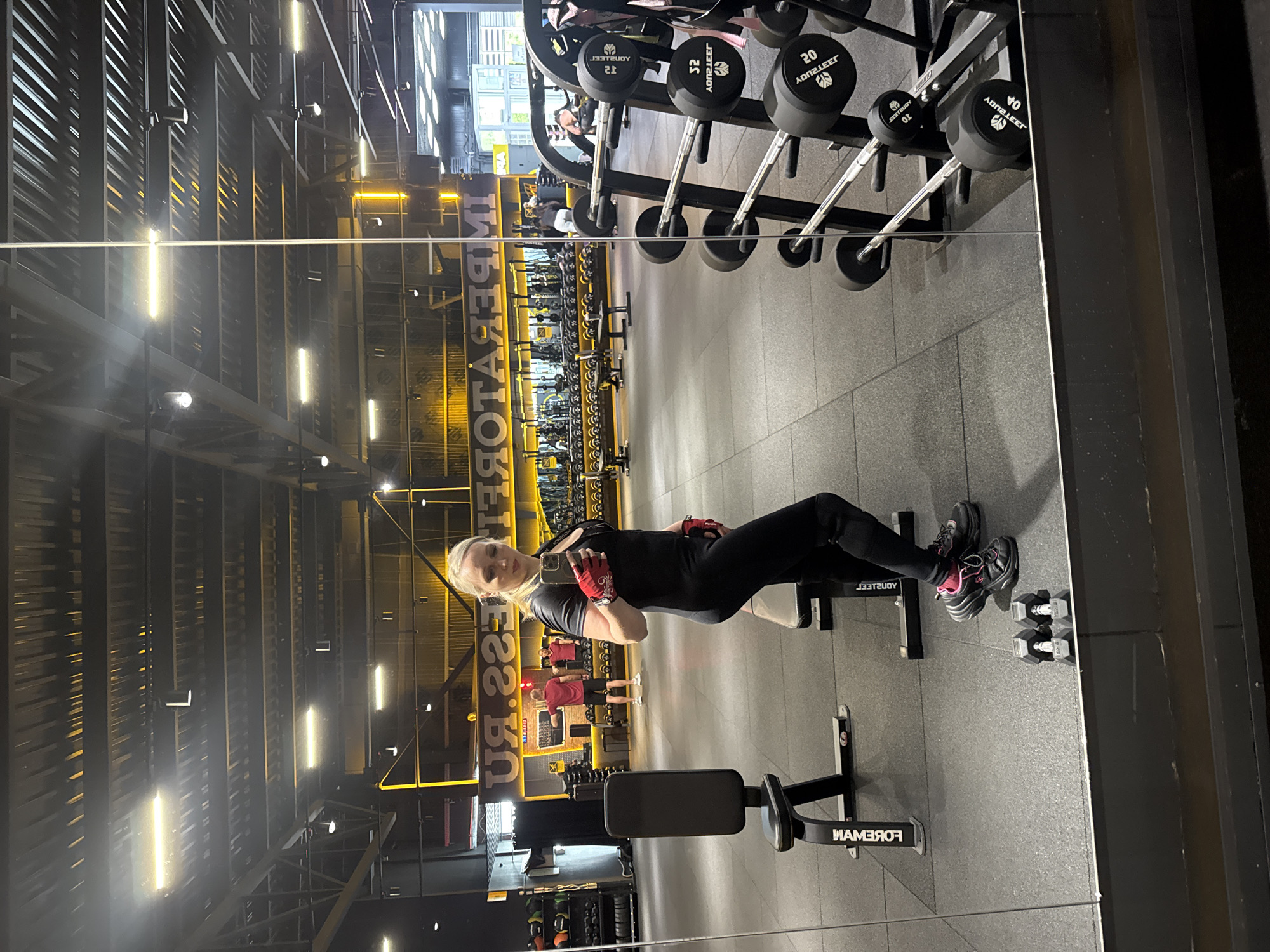 A blonde woman in black athletic wear and red gloves is taking a mirror selfie in a gym. She is standing in front of a large mirror that reflects the gym equipment behind her, including weight racks, benches, and dumbbells. In the reflection, two men are visible in the background, one wearing a red shirt and shorts, the other in a white t-shirt and shorts. The gym appears to be well-lit with overhead fluorescent lights and industrial-style lighting. A large sign with yellow lettering that appears to read "IMPERATOR.RU" is mounted on the wall behind the reflected figures. The floor is dark grey and textured, typical of a gym environment. The gym is located in Komsomolsk-on-Amur, Russia.
