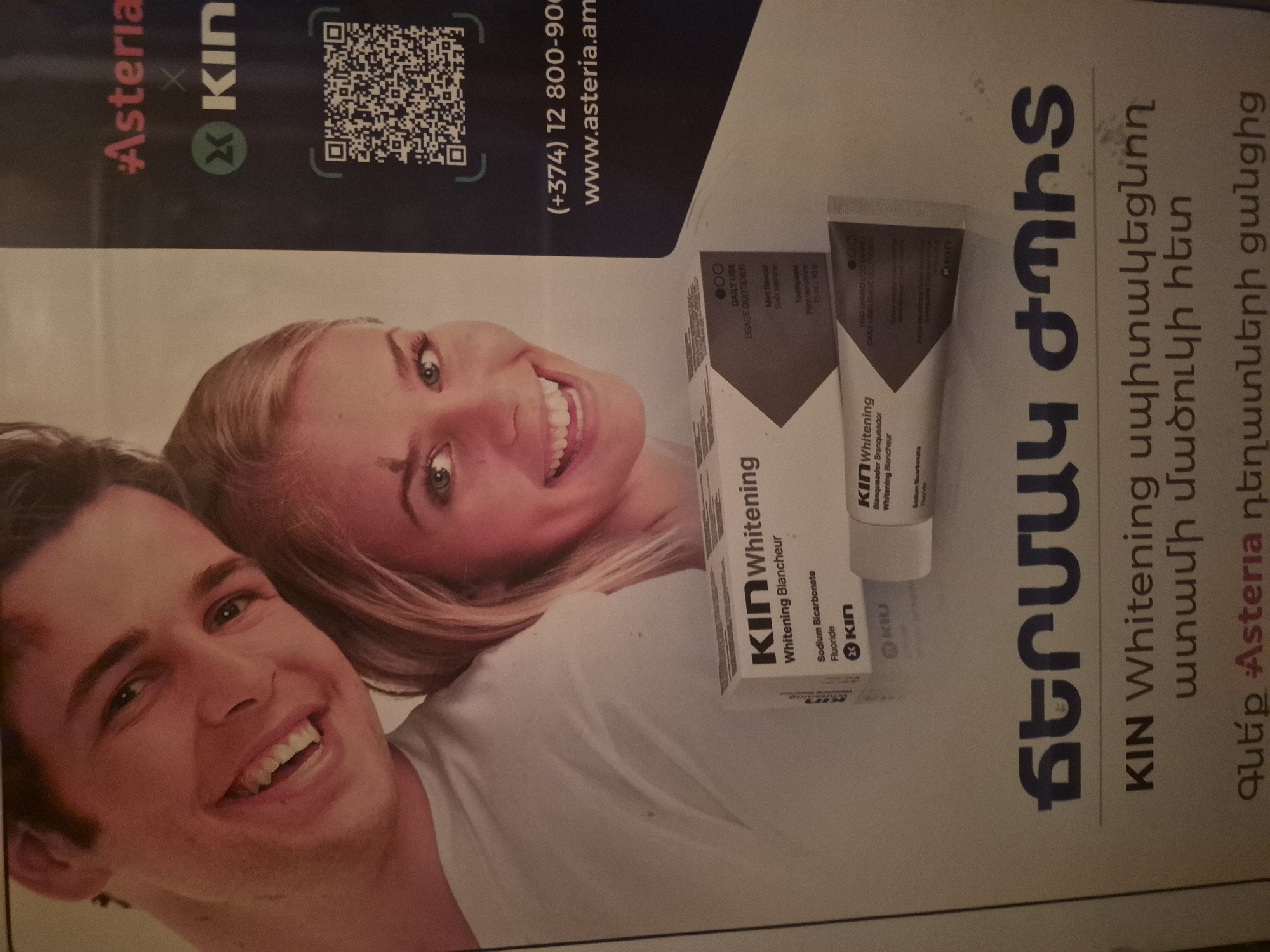 A promotional advertisement, likely a poster or large print, showcases two smiling individuals, a man and a woman, with bright white teeth. The woman is positioned in the upper part of the image, and the man below her, both looking towards the viewer with happy expressions. In the foreground, resting on the image of the woman's shoulder, are a box and tube of "KIN Whitening" toothpaste. The packaging details "Sodium Bicarbonate" and "Fluoride" as ingredients, and indicates "DAILY USE" and "Mint flavour" in English, French, and Spanish.
The background of the advertisement is dark blue, featuring several textual elements and a QR code. Branding includes "#Asteria" and "KIN" logos. Contact information is visible, stating "(+374) 12 800-900" and "www.asteria.am". The prominent Armenian text reads "ՃԵՐՄԱԿ ԺՊԻՏ" (WHITE SMILE), followed by "KIN Whitening սպիտակեցնող ատամի մածուկի հետ" (With KIN Whitening toothpaste), and "Գնե՛ք Asteria դեղատների ցանցից" (Buy from Asteria pharmacy network). The overall impression is a vibrant advertisement for dental hygiene products, encouraging purchases from Asteria pharmacies in Armenia.