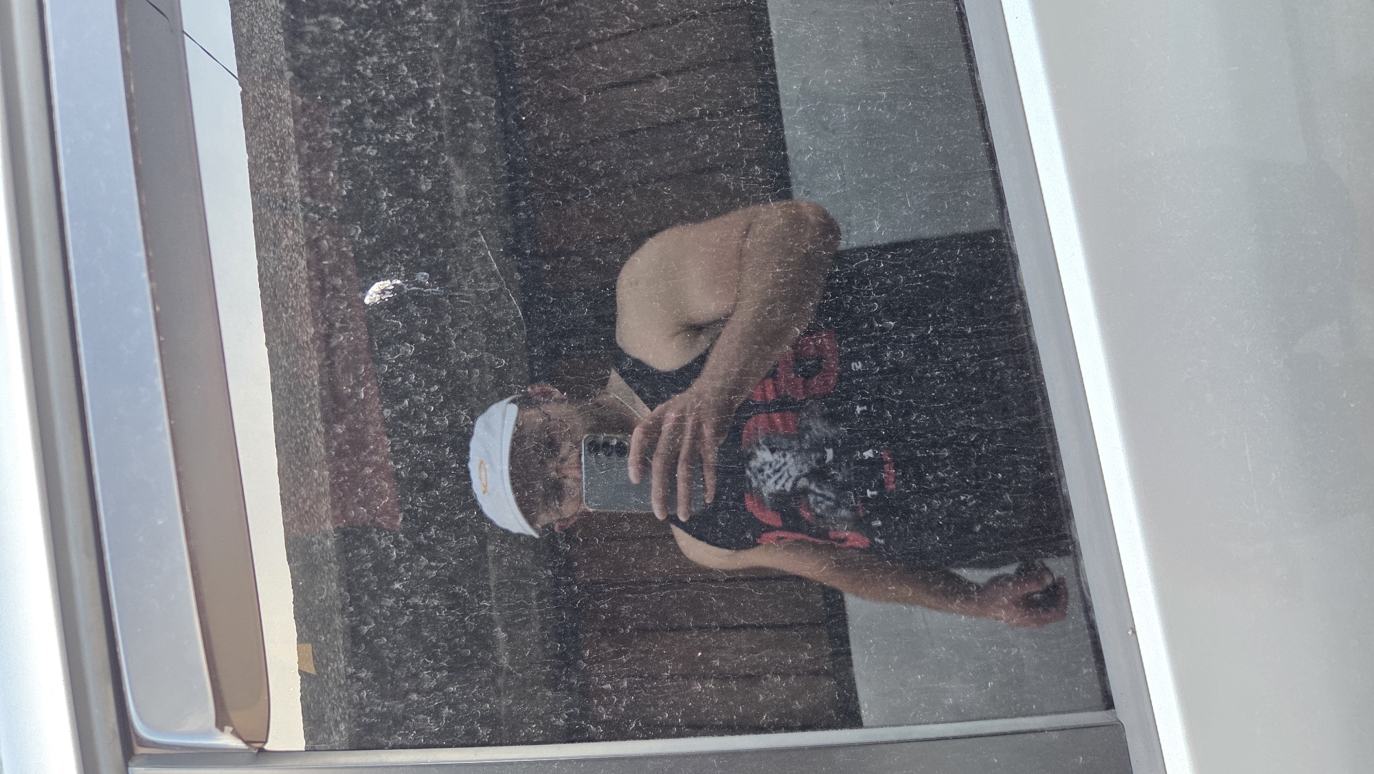 The image is a close-up, vertically oriented reflection in what appears to be a dirty window or mirror. The reflection shows a person from the chest up, taking a selfie with a smartphone. The person is wearing a white head covering with an orange logo, a black tank top with a red graphic design, and patterned shorts. The background of the reflection consists of weathered wooden planks, suggesting an outdoor setting. The overall lighting indicates it is daytime, possibly sunny due to the bright sky visible on the left side of the frame. The window or mirror is smudged and dirty, with visible dust particles and streaks. There is a silver metallic frame or edge visible on the left and right sides of the image, likely part of the window. The image appears to be taken in Bania, Bulgaria, though this information is not directly visible in the image itself.