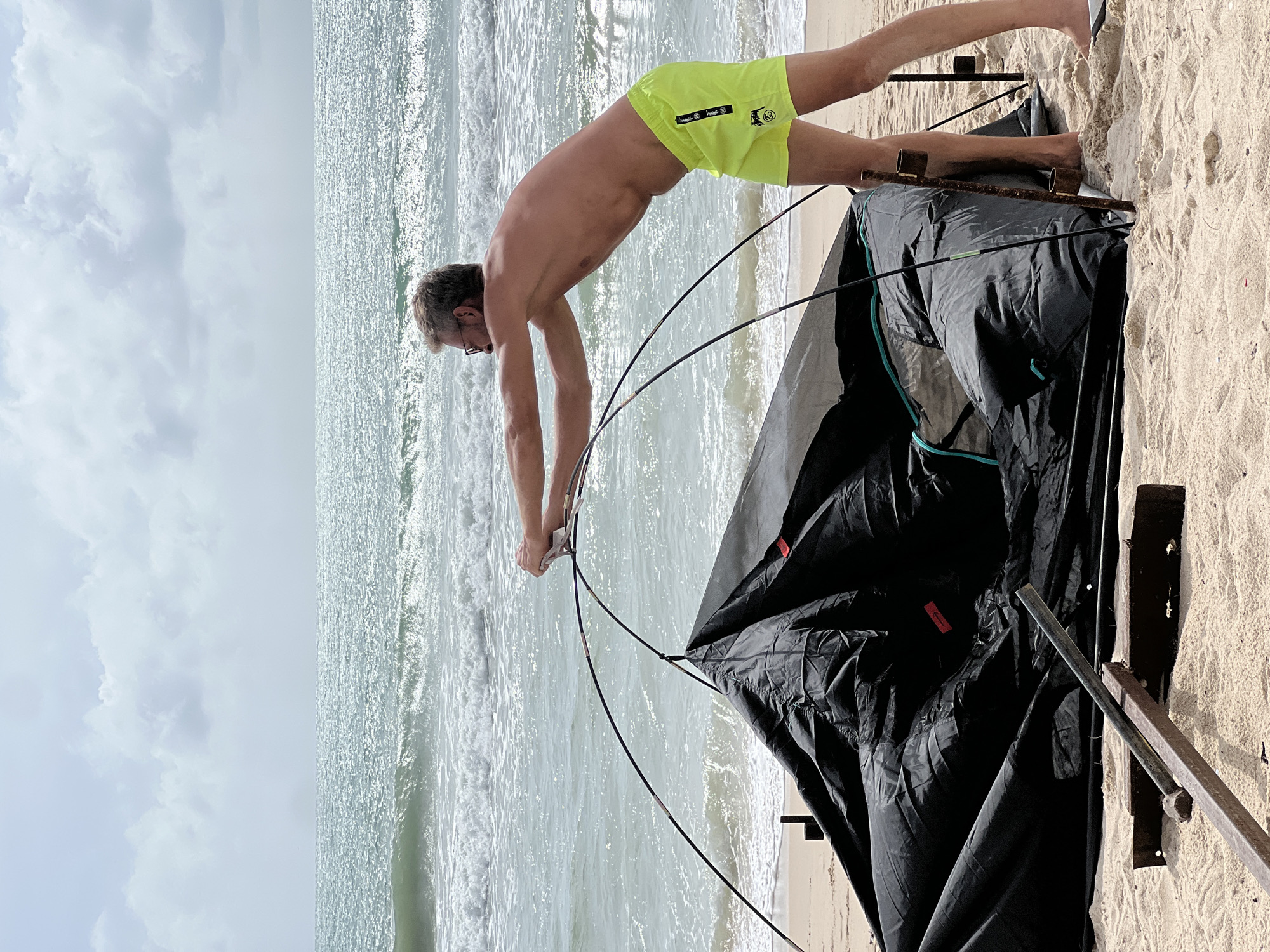 A shirtless man with glasses and blond hair, wearing bright yellow swim trunks, is on a sandy beach in Choeng Thale, Thailand, assembling a black tent. He is bending over, holding onto the tent poles, with his hands near the top of the tent. The tent is partially erected, with its fabric spread out on the sand. The ocean with gentle waves is visible in the background, under a cloudy sky. In the foreground, there are some pieces of metal and wooden debris on the sand.