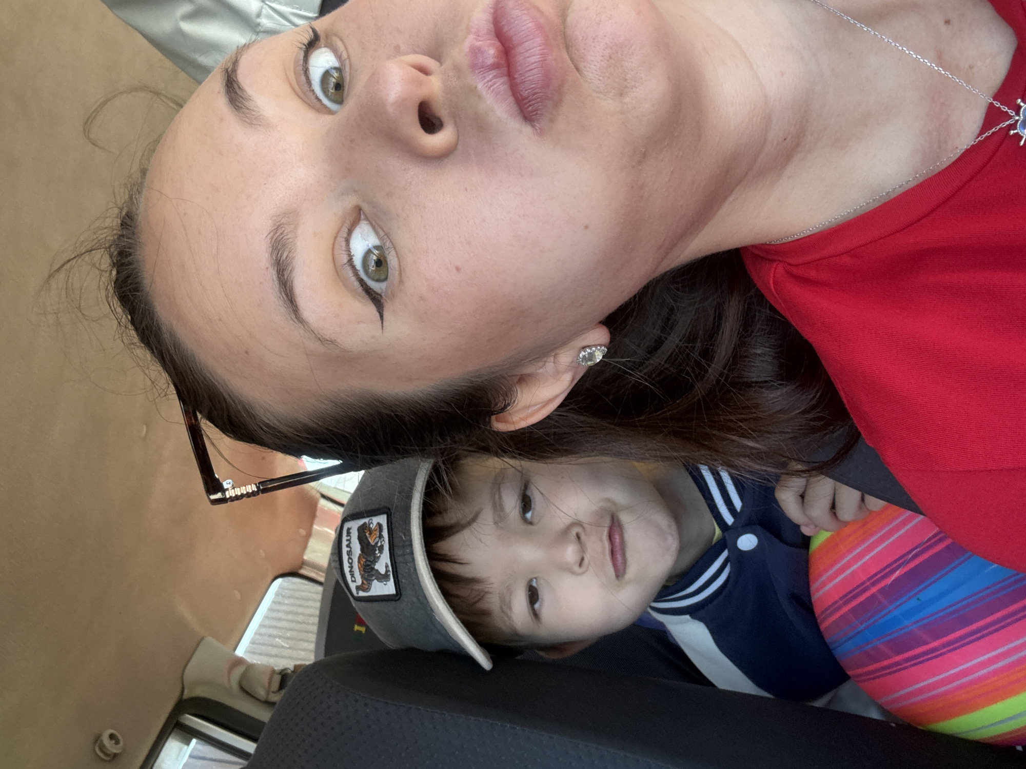 This is a close-up, selfie-style photograph taken inside a vehicle. The image features two individuals, an adult female and a young child. The adult female is in the foreground on the right, her face taking up a significant portion of the frame. She is looking directly at the camera with wide, surprised eyes and a pouty expression. She is wearing a red shirt and has on winged eyeliner and a diamond stud earring. The young child is seated behind and to the left of the woman. He is wearing a baseball cap with a dinosaur patch and a blue and white jacket. The child has a somewhat sullen or unimpressed expression. A brightly colored, striped ball is visible in the lower left portion of the image, partially obscuring the child. The interior of the car is visible, including the headliner and a car seat. The lighting suggests it is daytime. The photograph was taken at the Bely Yar cemetery, located in Bely Yar, Russia.