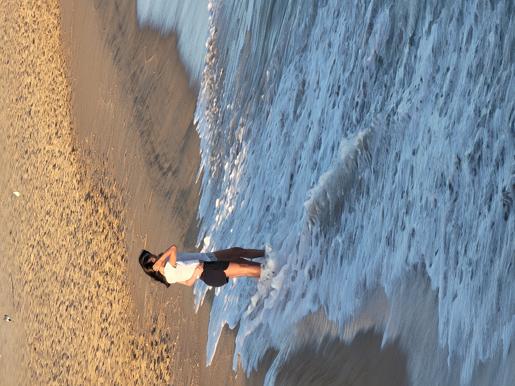 This media file captures a tranquil scene at the beach in Quarteira, Portugal. The image is oriented vertically, with the shoreline running diagonally from the bottom left to the top right. A young couple is the central focus, standing close together in the shallow surf. The woman, on the left, wears a white t-shirt and black shorts, with her arm around the man. The man, on the right, is dressed in darker clothing and has his arm around her waist. They appear to be embracing, their bodies angled towards each other, suggesting a moment of intimacy or connection.
The waves are gently rolling in, creating a frothy, white foam that surrounds their legs and feet. The water is a muted blue, reflecting the soft light of either dawn or dusk, indicated by the warm, golden glow on the sand. The sand itself is a textured expanse of light brown, marked with subtle ripples and scattered with small pebbles. To the left of the couple, a solitary bird walks on the wet sand, adding a touch of natural life to the scene.
The overall mood is peaceful and serene, enhanced by the soft lighting and the gentle motion of the water. The setting is clearly a beach, with the distinct textures of sand and seafoam, and the warm light suggesting a pleasant climate. There is no visible text in the image.