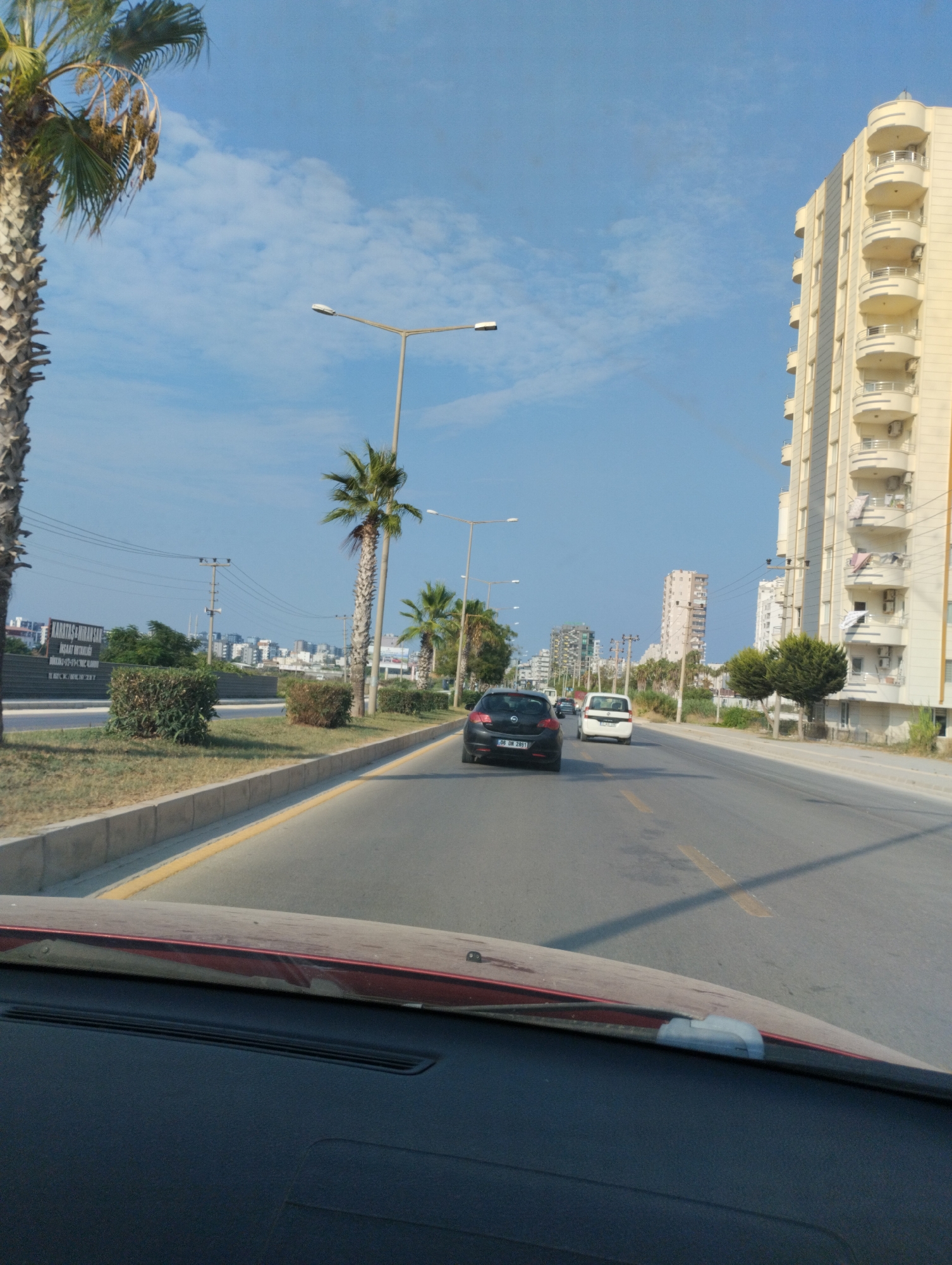 A first-person perspective shot from inside a car driving on a multi-lane road in Mezitli, Turkey. The sky is blue with scattered clouds. On the right side of the road is a tall, cream-colored apartment building with many balconies. On the left side of the road are palm trees, bushes, and a sign that reads "KARATAŞ İNŞAAT SAN". Several cars are visible on the road, including a black car with a license plate that reads "06 DM 2891" and a white minivan. In the distance, other buildings and palm trees can be seen. The car's dashboard is visible in the foreground.