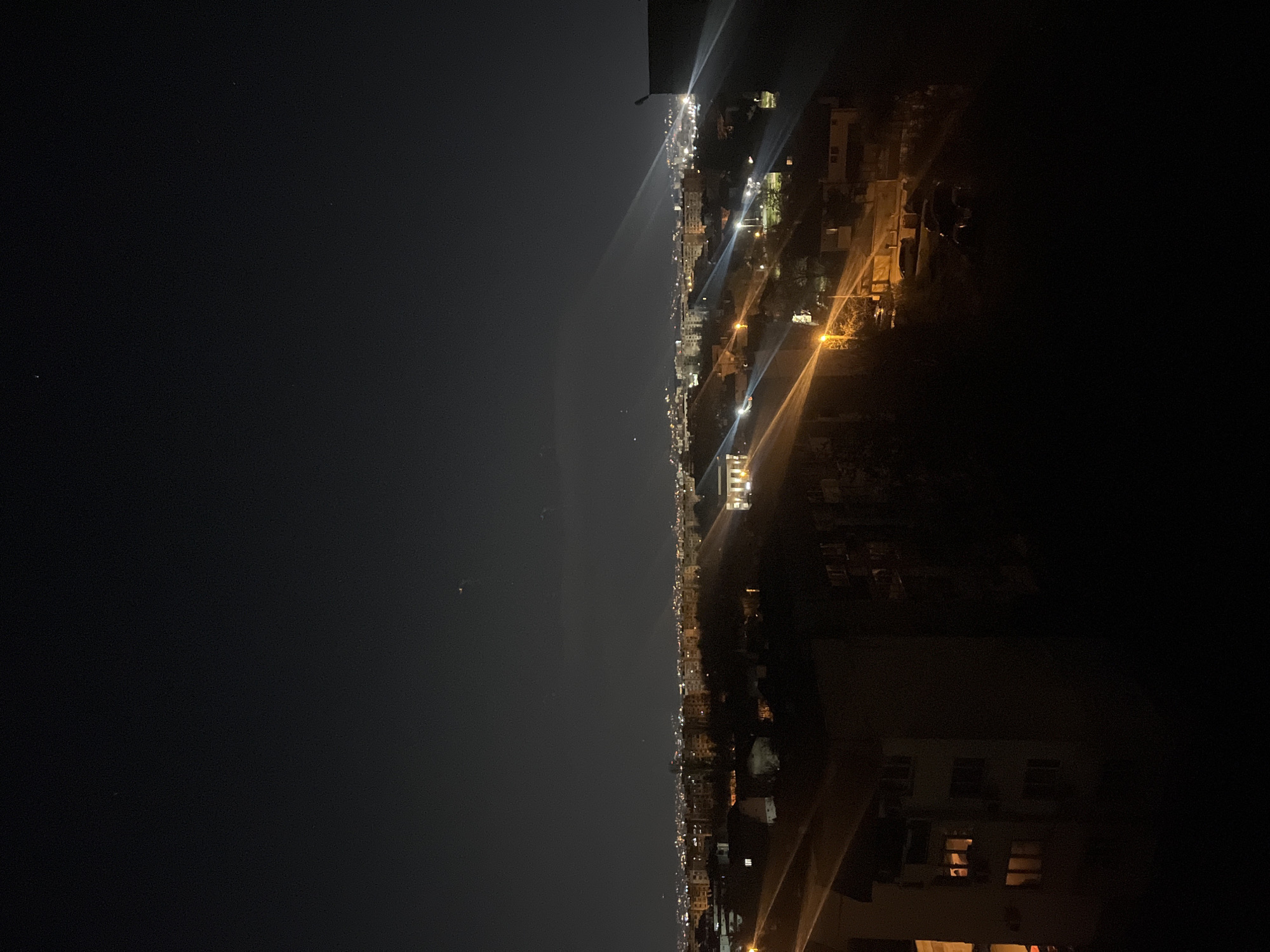 The image shows a nighttime cityscape, likely taken from a high vantage point in Sofia, Bulgaria. The scene is dominated by the city lights stretching into the distance and a dark night sky above. On the right, a tall building is visible, with several illuminated windows. The streetlights and building lights indicate the presence of residential and commercial areas. The night sky on the left appears clear, with a few stars faintly visible. The overall atmosphere is calm and urban, with a focus on the artificial illumination of the city at night.