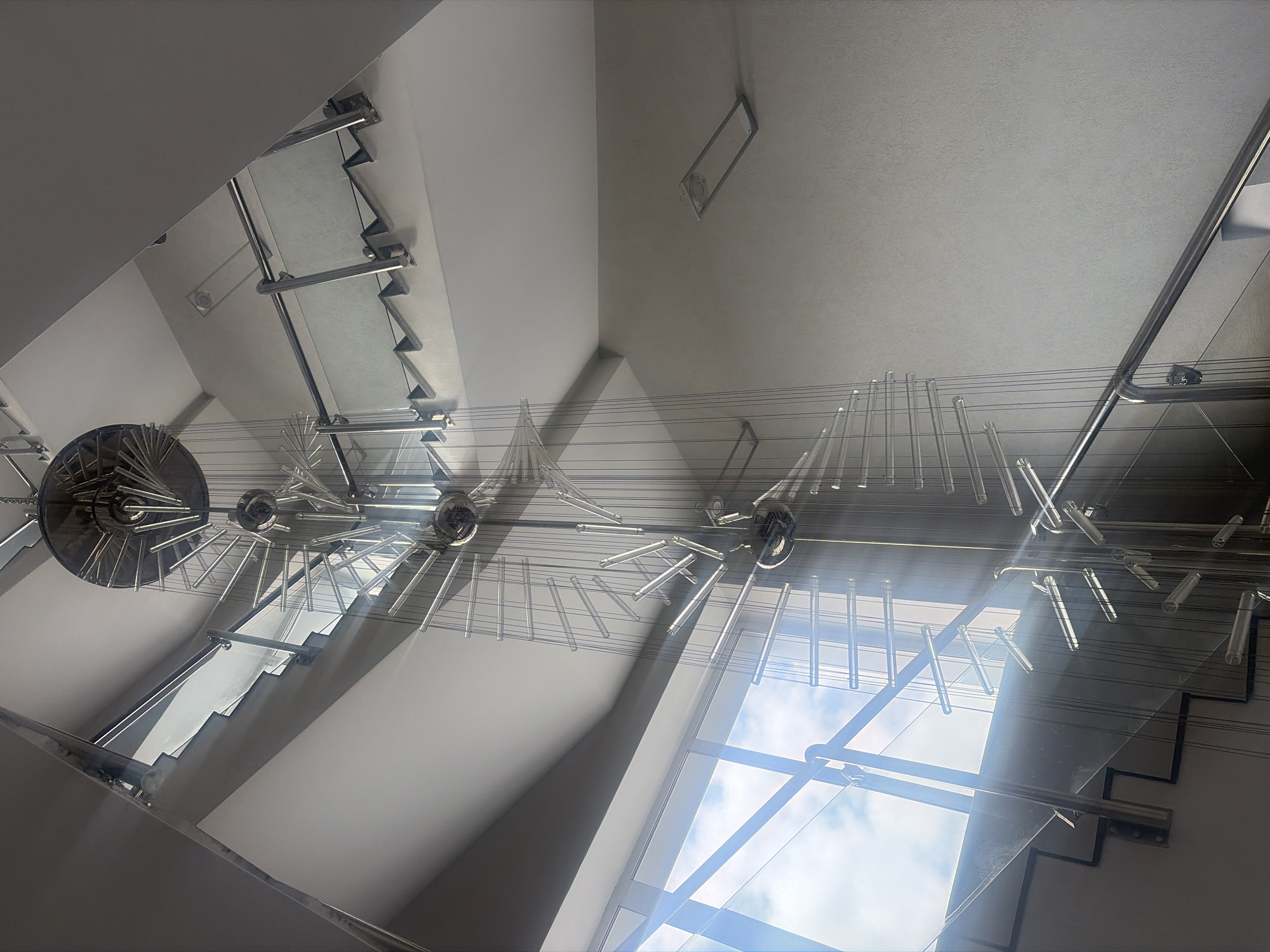A low-angle, high-contrast shot shows the interior of a building in Saint Petersburg, Russia. The main features are a staircase and a large, ornate lighting fixture suspended from the ceiling. The staircase is modern, with glass panels and silver-colored metal supports. The lighting fixture is complex, featuring multiple circular elements and dangling glass tubes or rods. Bright light streams in from a window, creating strong shadows and highlights on the staircase and other architectural elements. The walls and ceiling are a neutral, light color, adding to the brightness of the scene.