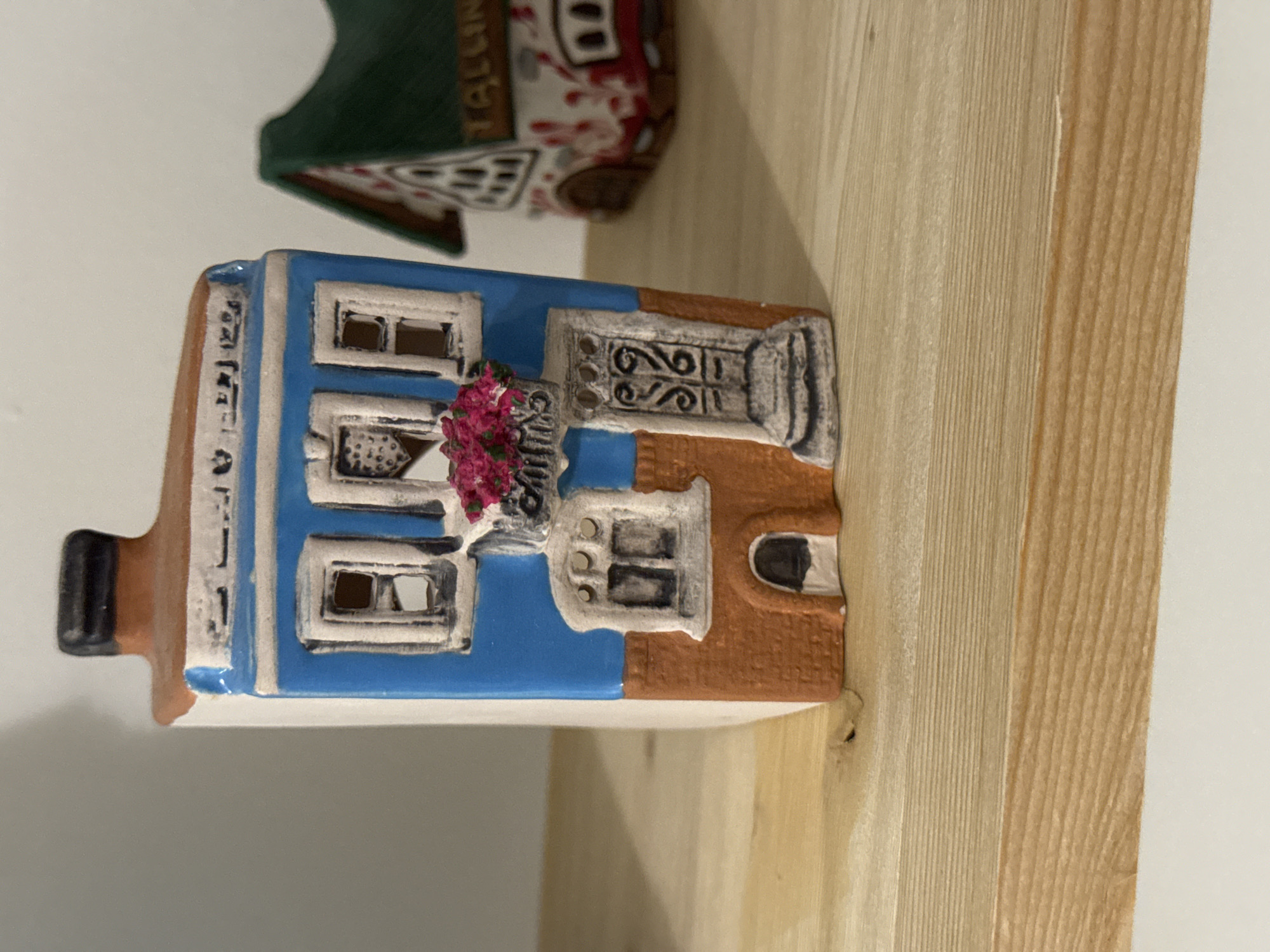 The image shows a ceramic miniature of a Dutch canal house placed on a wooden shelf. The house is predominantly blue, with white window frames and a red flower box beneath one of the windows. Part of another ceramic building with a green roof is visible in the upper left corner. The shelf is made of light-colored wood with a visible grain. There is no indication that this is located in Izhevsk, Russia.