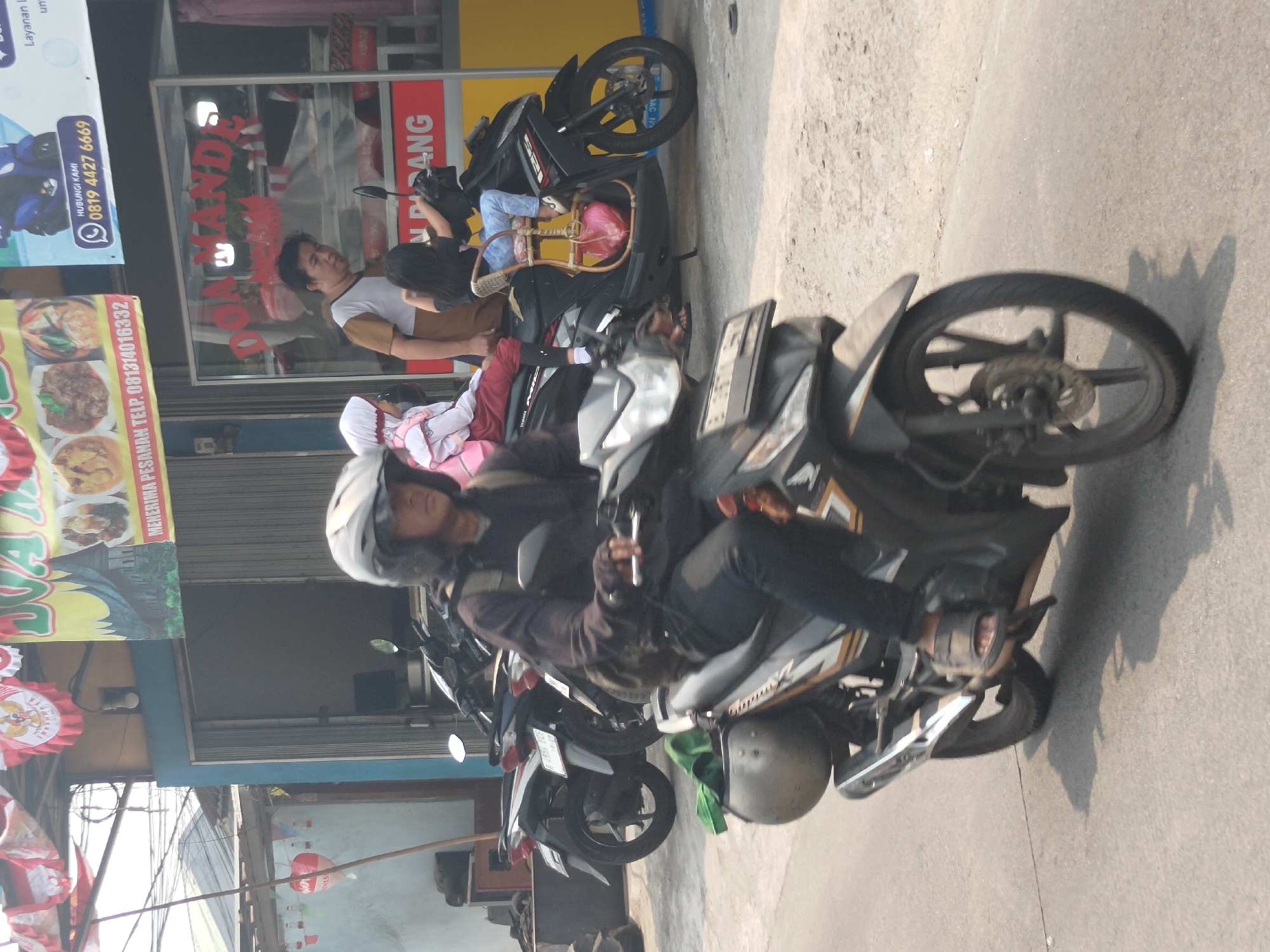 In Depok, Indonesia, a man wearing a helmet and a dark jacket is riding a motorcycle. He is wearing black pants and sandals. The motorcycle is black and gold. A woman wearing a headscarf and a light pink jacket, and a young girl in a white and pink school uniform are sitting on the back of the motorcycle. The motorcycle is in motion on a paved road. In the background, there is a building with a blue and yellow facade and a sign that says "DOA MANDE". There are also other motorcycles parked in front of the building. The weather appears to be sunny.