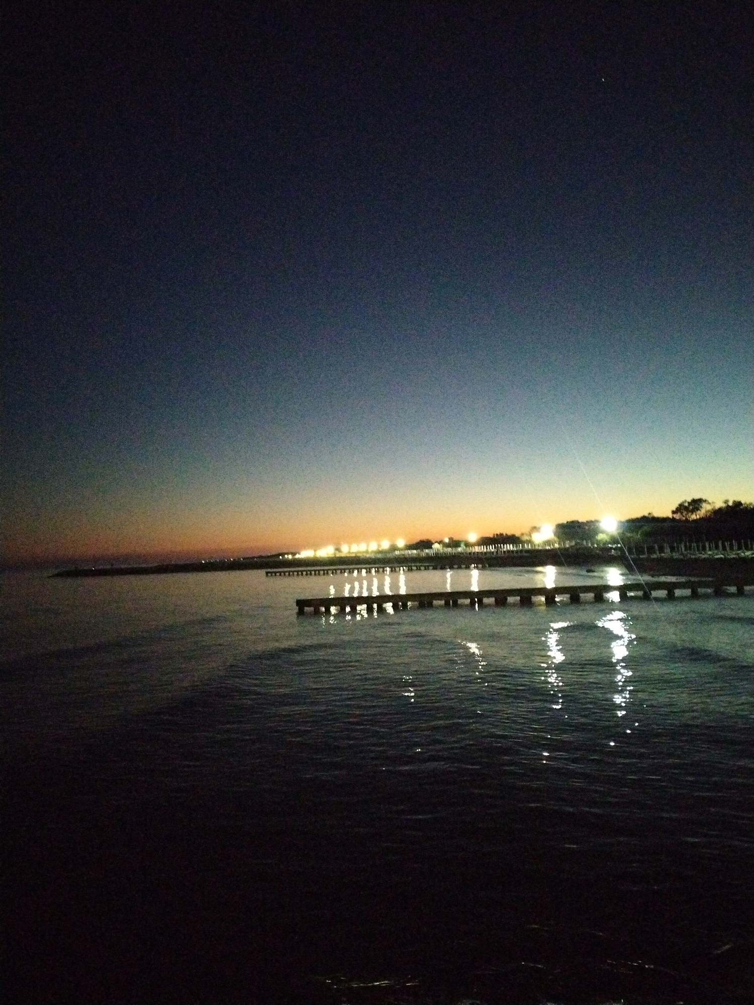 The image depicts a twilight scene at the Caorle seafront, Italy. The sky transitions from a deep indigo at the top to a soft orange and yellow hue near the horizon, indicating either sunrise or sunset. A faint, crescent-shaped celestial body is visible in the upper right quadrant of the sky.
The foreground is dominated by dark, rippling water, reflecting the ambient light from the shore. Two piers extend into the water. The closer pier is a dark, wooden structure, partially submerged and its pilings illuminated by the reflection of lights from the shore. The further pier is a longer, darker structure that stretches towards the left of the frame, with a line of lights visible along its length.
Along the distant shoreline, a row of lights, likely streetlights or lights from buildings, illuminates the tree line. The overall atmosphere is serene and quiet, with no human presence explicitly visible in the image. The scene is characteristic of a peaceful evening or early morning by the Adriatic Sea in Caorle.