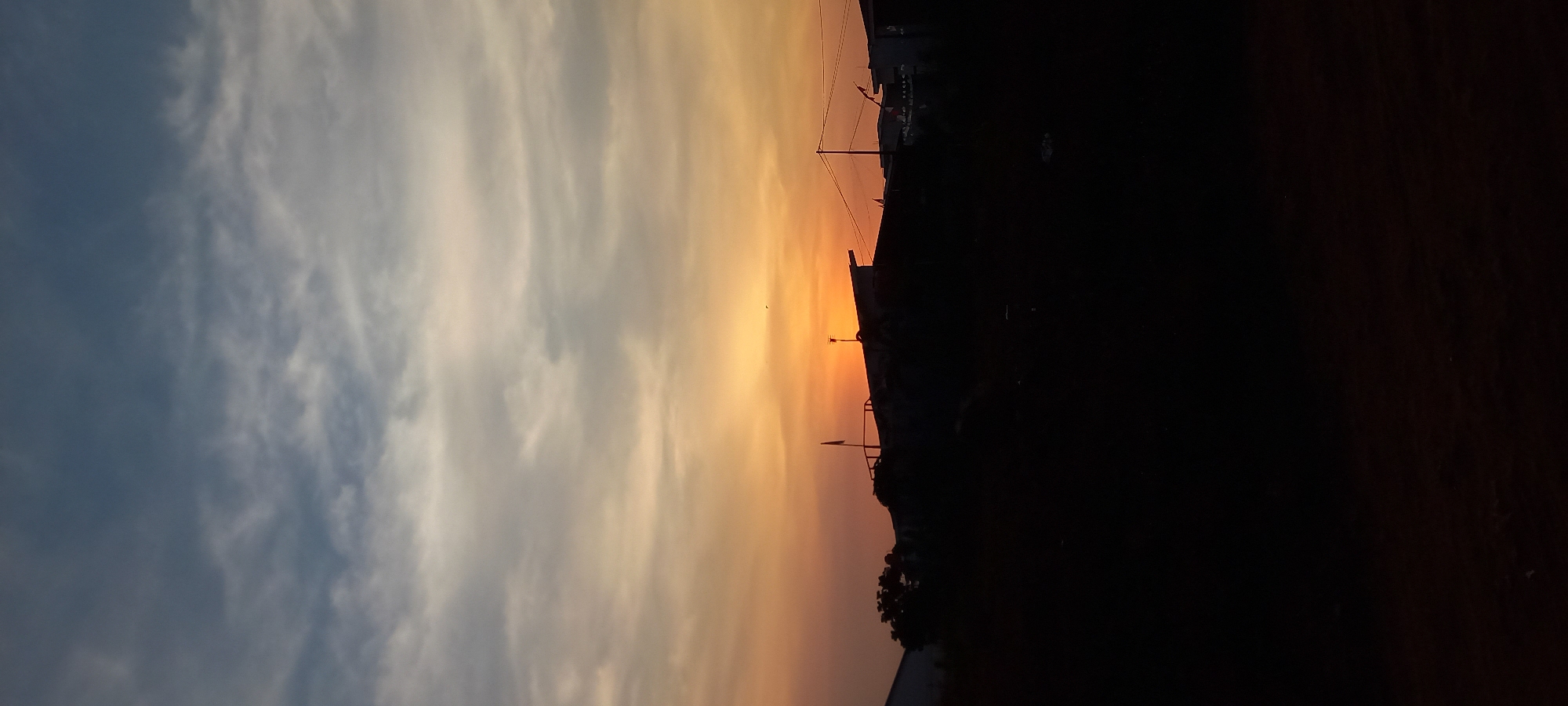 The image shows a silhouette of buildings and trees against a sunset sky. The sky is a gradient of colors, from a deep blue at the top to a warm orange and yellow near the horizon. There are clouds scattered across the sky, with some appearing wispy and others more dense. In the foreground, the silhouettes of buildings, trees, and what appear to be some flags are visible against the bright sky. The lower part of the image is very dark, obscuring details of the ground. The scene suggests it was taken in Loa Lepu, Indonesia, during sunset.