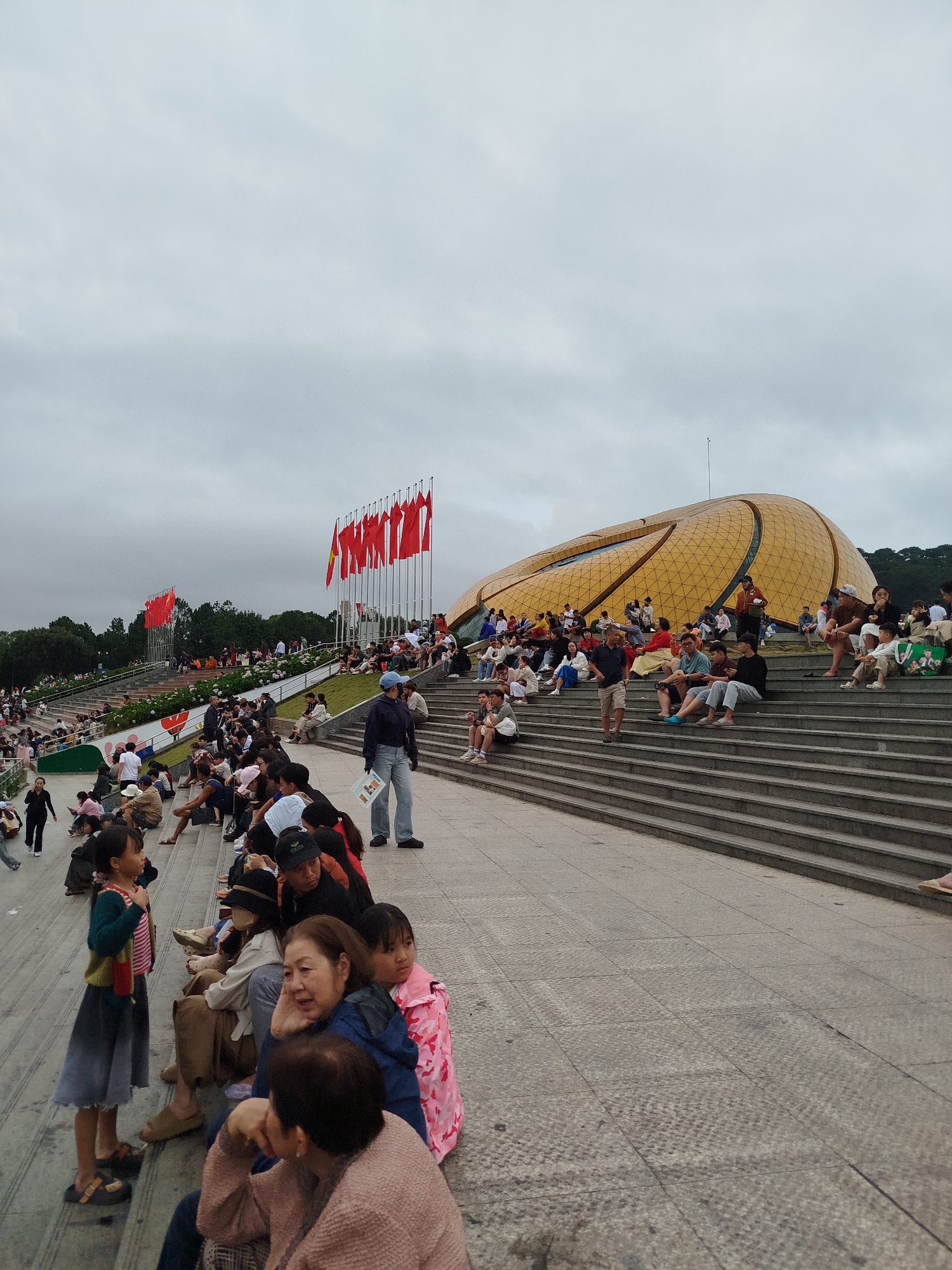 The image depicts a gathering of people seated on large concrete steps leading up to a distinctive golden, geodesic dome-shaped building. The building appears to be a modern architectural structure. In the background, several red flags with yellow stars are displayed on tall poles, suggesting a national or patriotic event. The sky is overcast and appears to be a light grey. The people are dressed in casual clothing, and many are sitting, talking, or observing. Some are children, and others appear to be adults and elderly individuals. The scene suggests an outdoor public event or gathering. This location is identifiable as the Da Lat Flower Park, Vietnam, specifically the area in front of the city's exhibition hall, which is known for its unique golden dome.