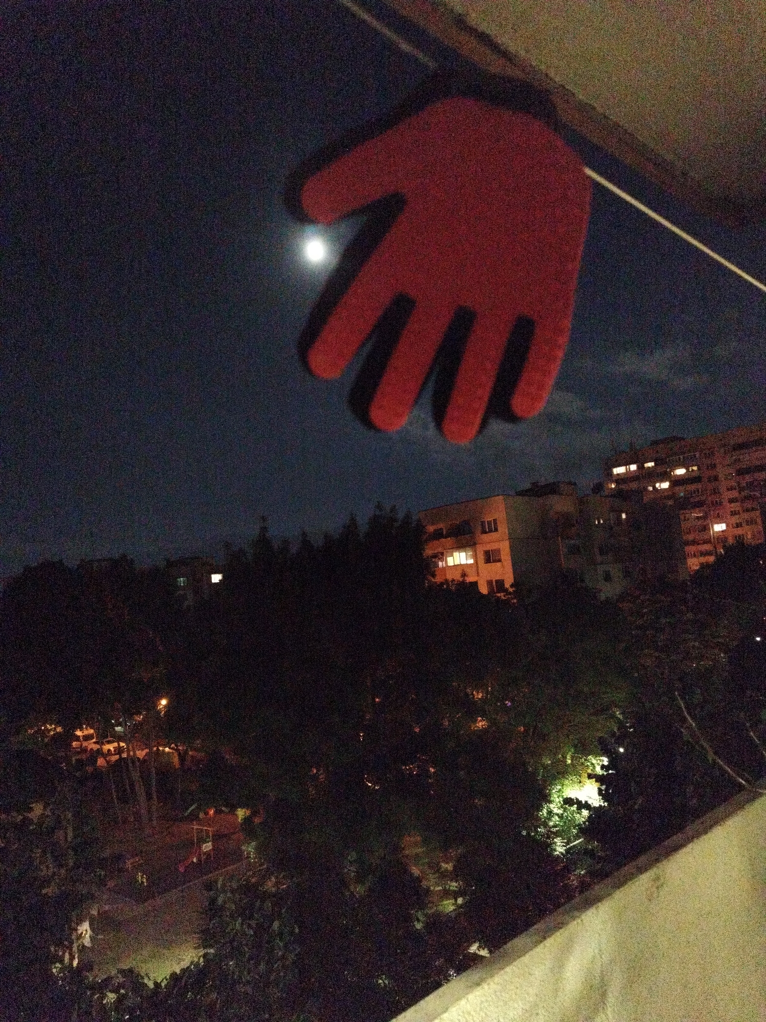 The image shows a night view, likely taken from a balcony in Sofia, Bulgaria, given the typical urban landscape. A red, cut-out hand shape is suspended in the foreground against the night sky where the moon is faintly visible. Below, the skyline is dominated by buildings with illuminated windows. Dense foliage, possibly trees, obscures the lower part of the view. In the dimly lit area, a playground can be recognized in the bottom left side of the view. A concrete ledge of a balcony can be seen at the bottom right corner of the image. No people are visible.